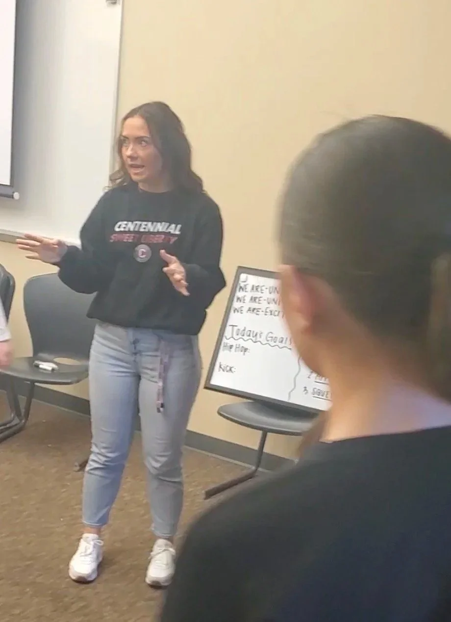 A woman standing in front of a classroom giving a presentation, with a whiteboard nearby and students seated and listening.