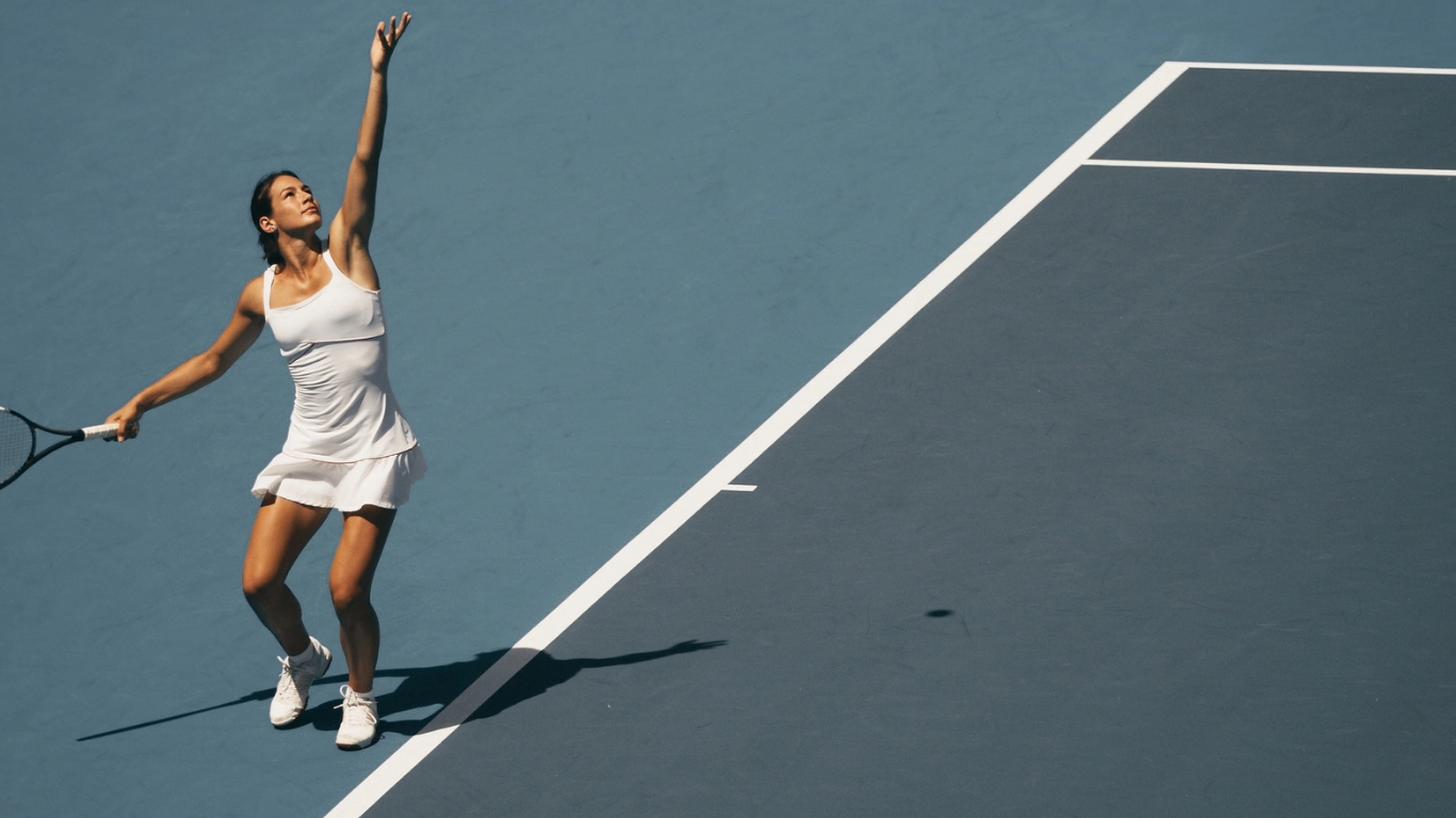 A woman in a white sleeveless tennis dress holding a tennis racket and a tennis ball on a tennis court.