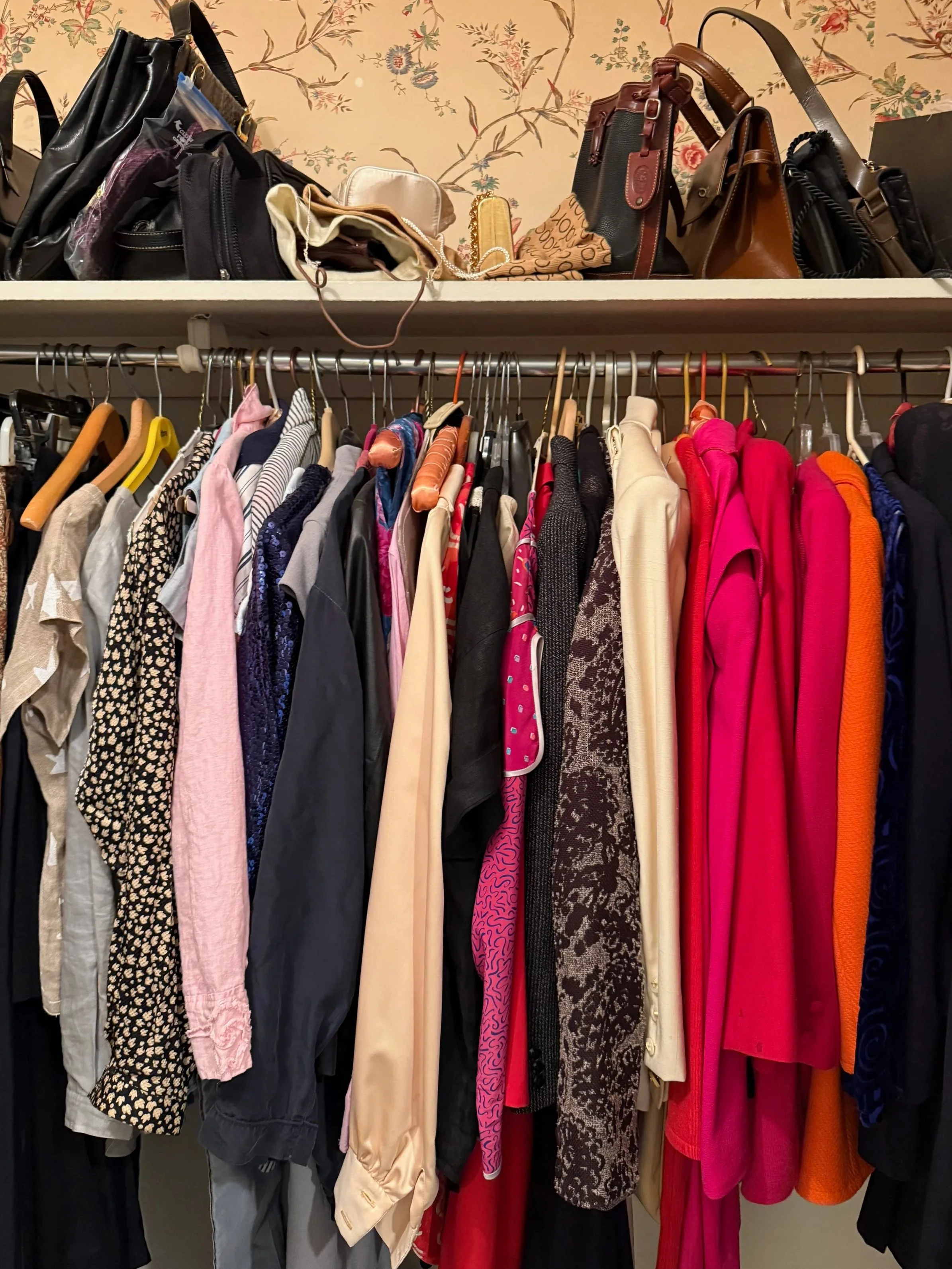 Clothes hanging on a rack including tops, jackets, and dresses in various colors and patterns, with handbags and purses on a shelf above, all against a floral wallpaper background.