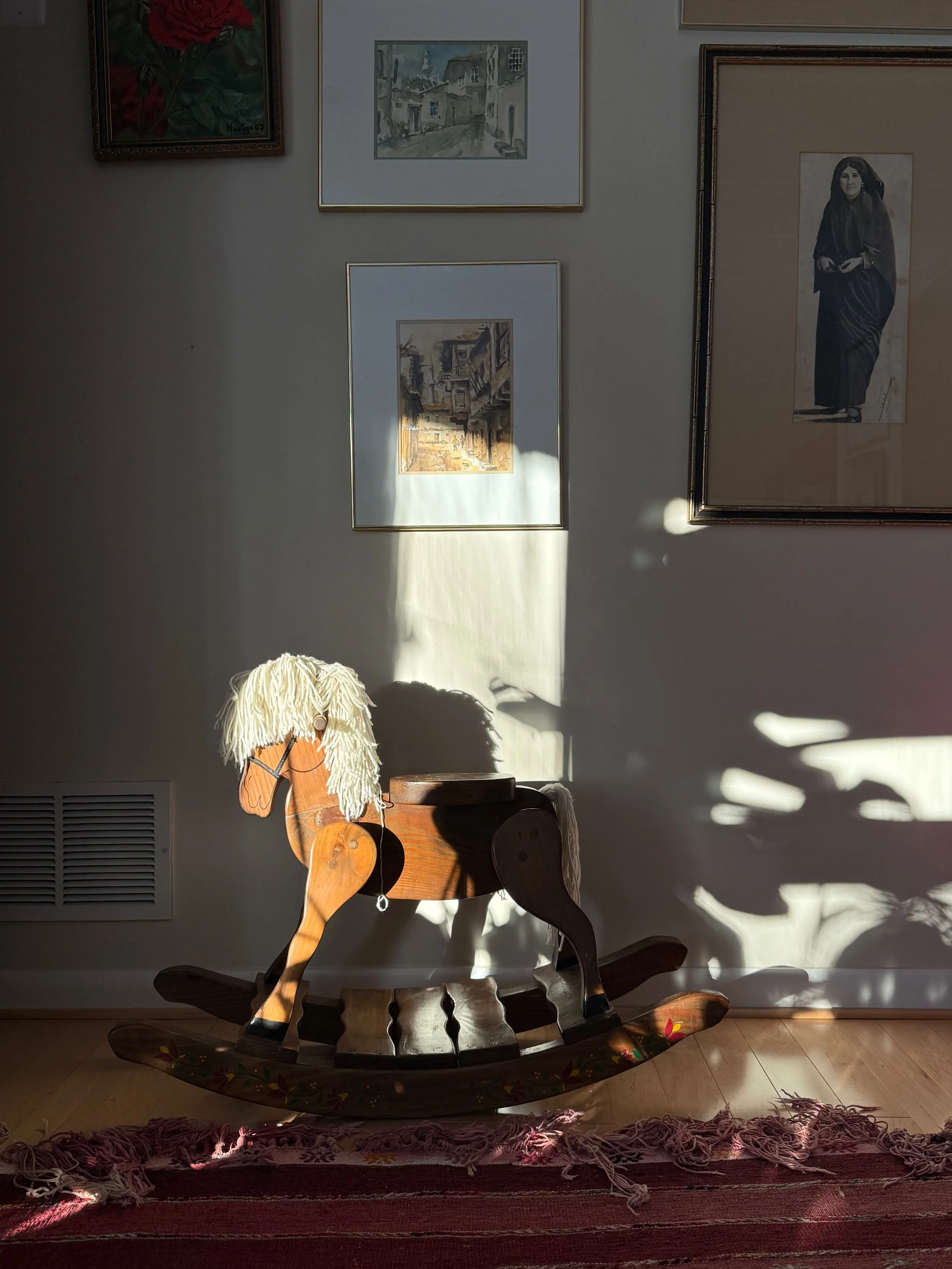 A wooden rocking horse with a white yarn mane, placed on a doormat in front of a wall with framed artwork, with sunlight casting shadows on the wall.