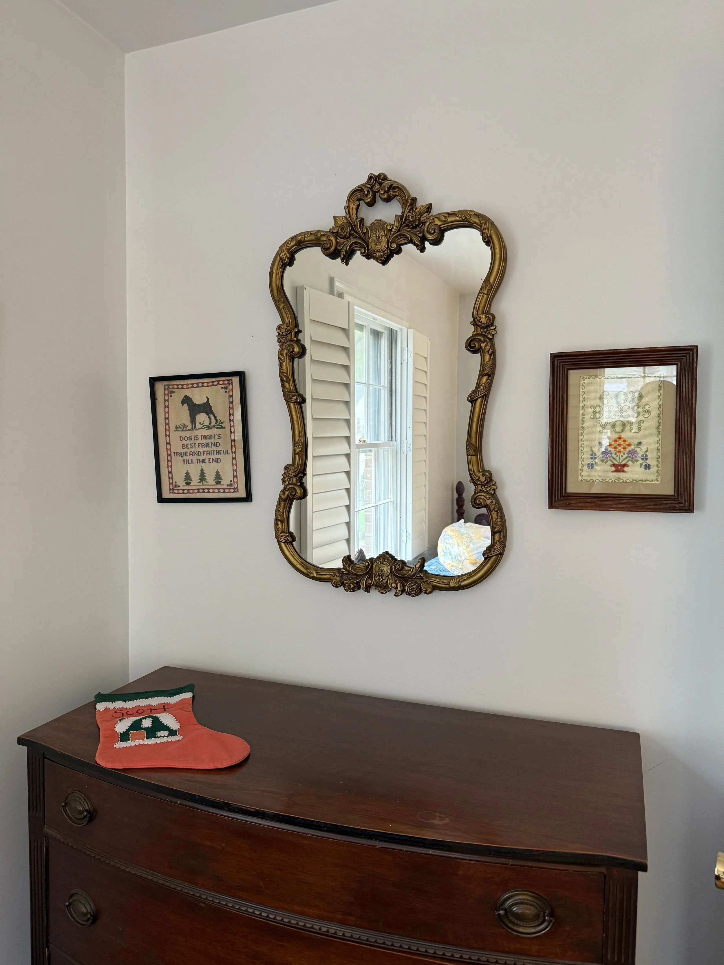A wooden dresser with a decorative Christmas stocking on top, a large ornate framed mirror hanging above, and two framed cross-stitch artworks on the white wall.