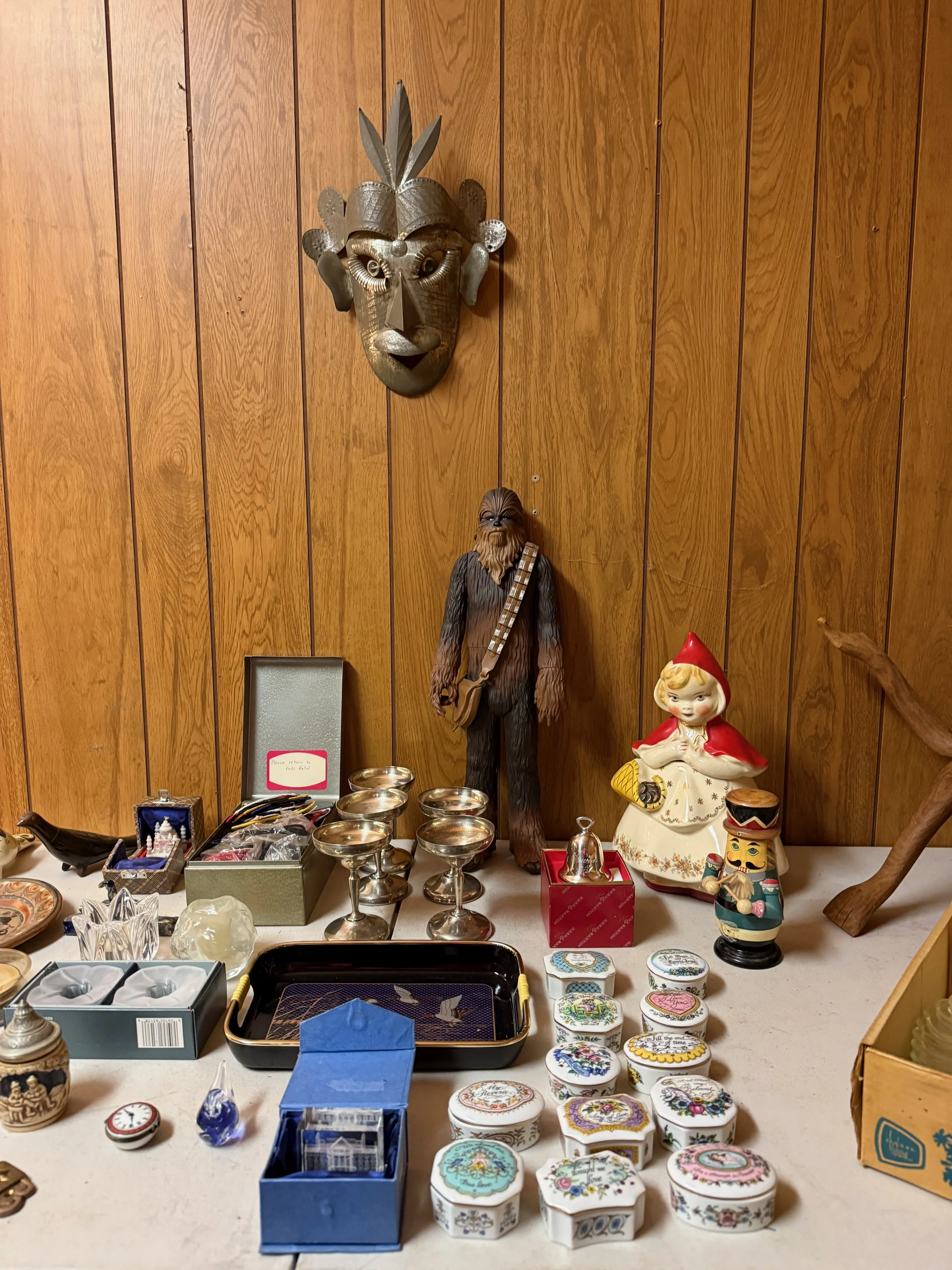A collection of vintage and decorative items displayed on a table against a wood-paneled wall, including a large tribal mask, a Chewbacca figurine from Star Wars, several decorative boxes, glassware, and figurines.