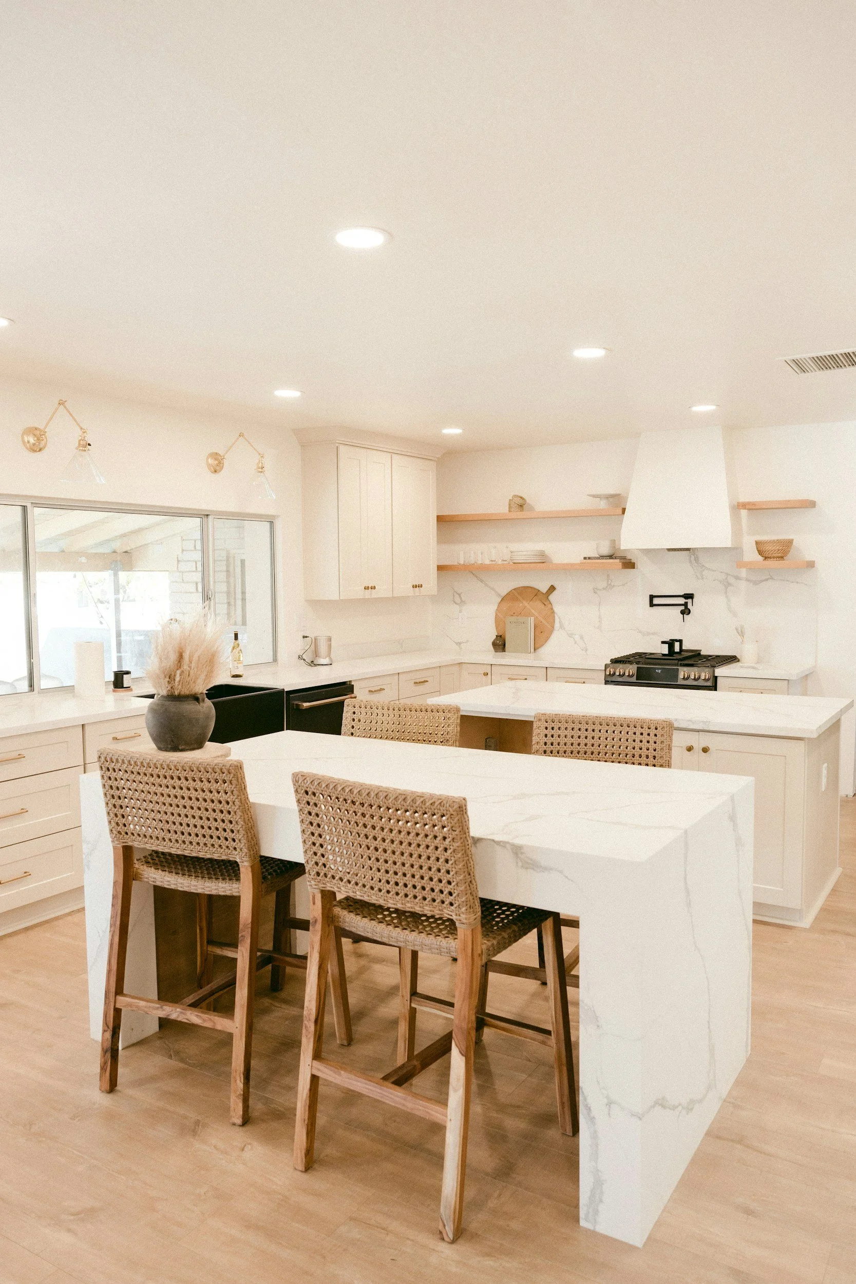 Bright, modern kitchen with white marble countertops, beige bar stools, and open wooden shelves on white walls, featuring a black stove, white cabinets, and a large window.