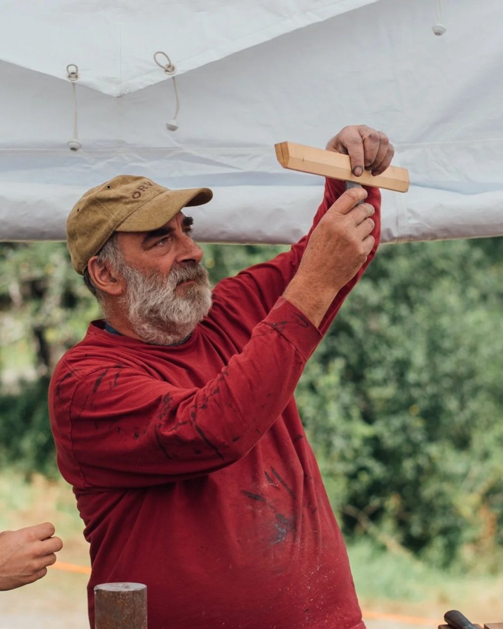 An older man with a gray beard wearing a tan cap and red shirt is hammering a nail into a wooden piece under a white canopy outdoors. There is greenery in the background.