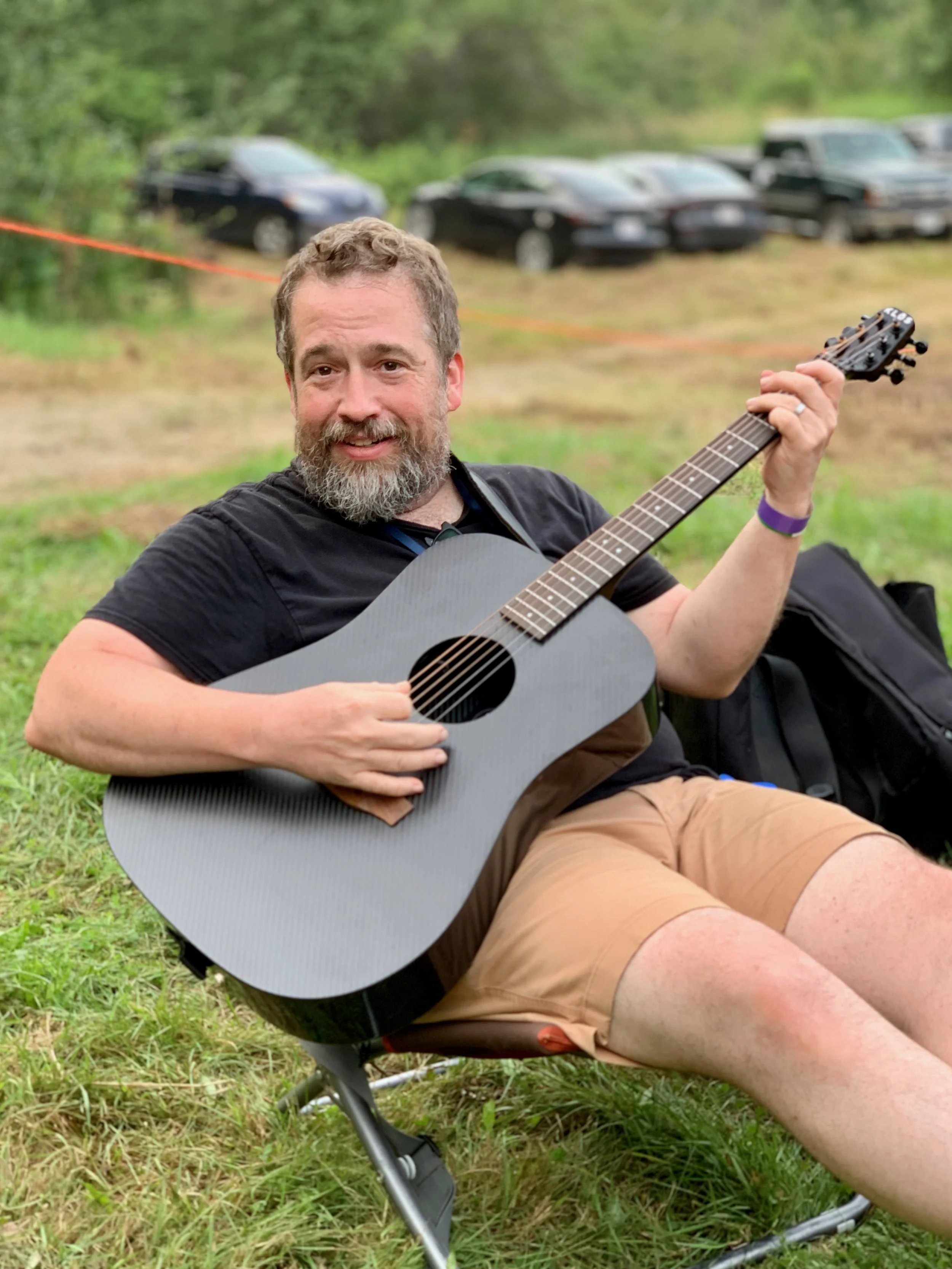 A man with a gray beard and short hair smiling while playing an acoustic guitar outdoors on a grassy area, with several parked cars and trees in the background.