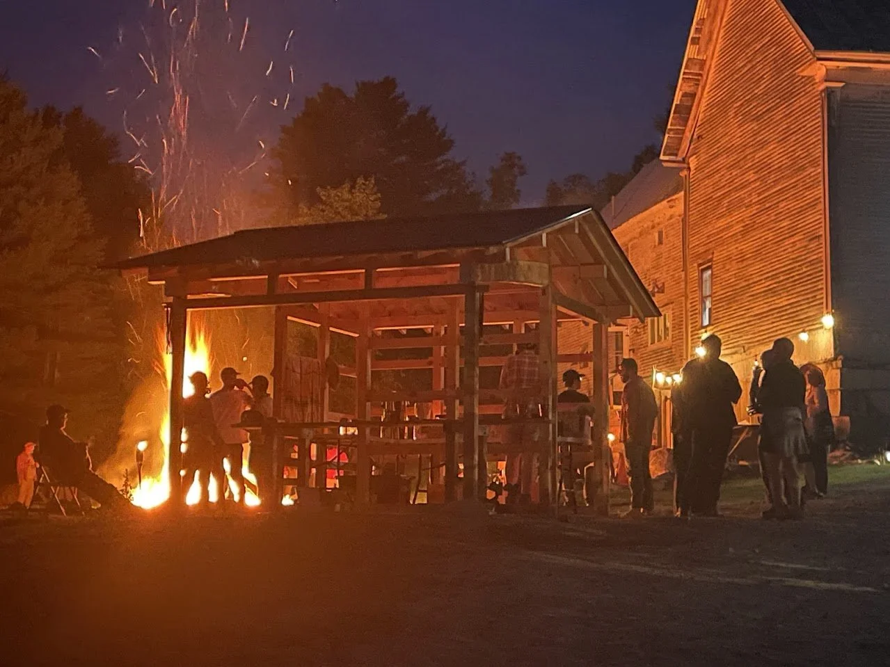 People gathered around a bonfire at night outside a wooden building. Fireworks are exploding in the sky above, casting light and shadows on the scene.
