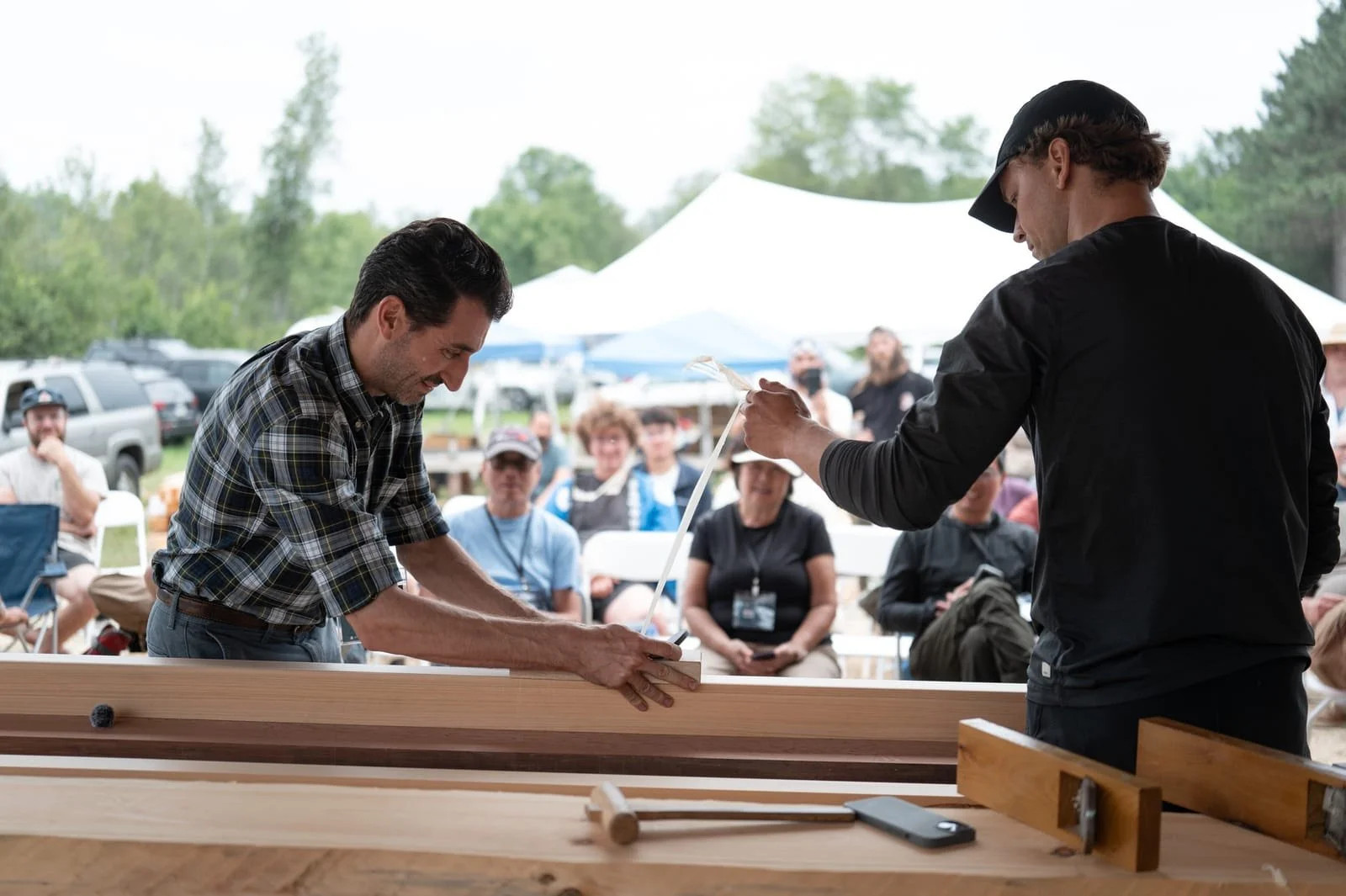 Two men give a woodworking demonstration at an outdoor event with an audience watching, tents, and trees in the background.