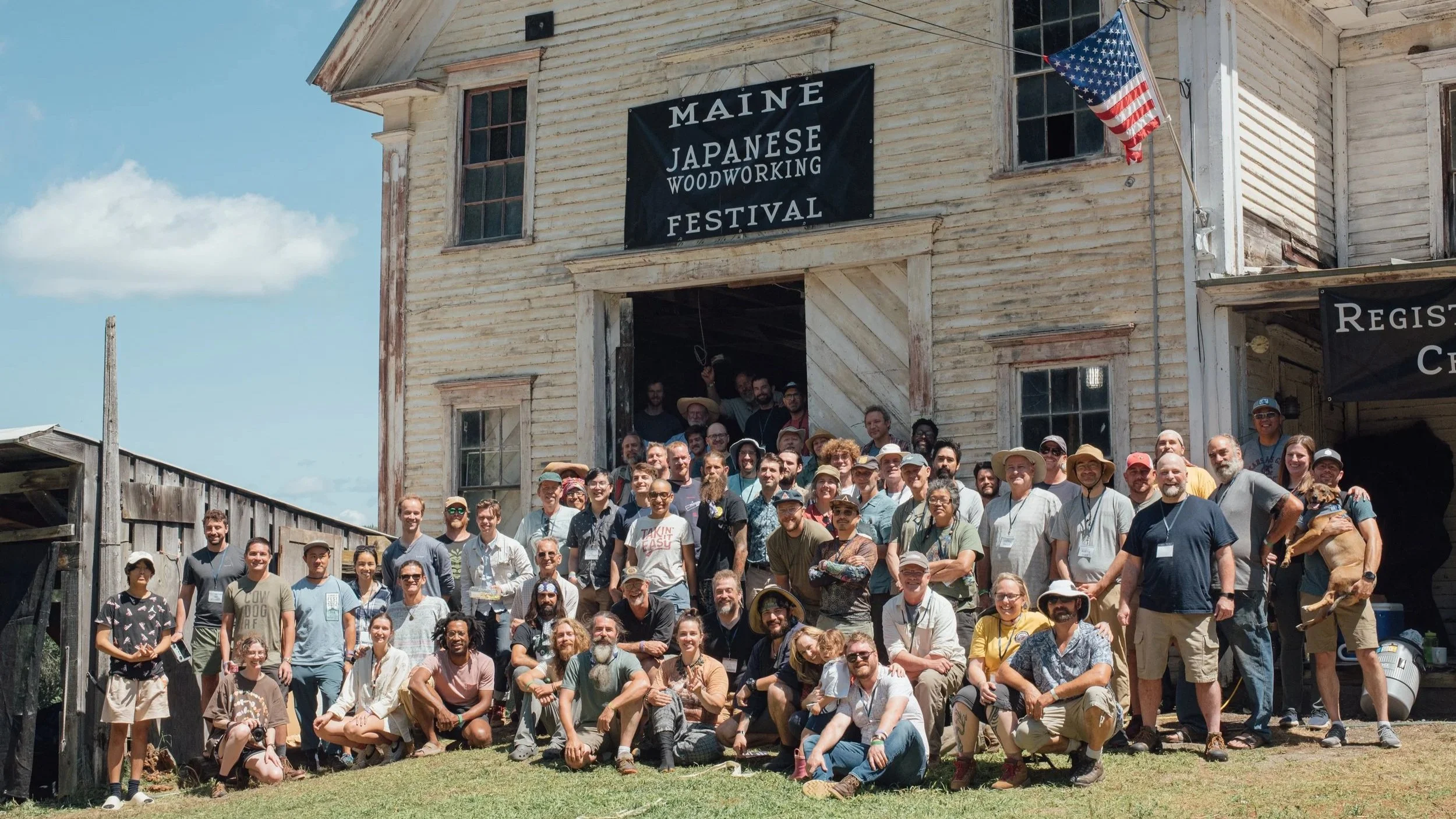 A large group of people posing in front of an old wooden building at the Maine Japanese Woodworking Festival. The building has a sign and American flag, with some people inside the building visible through the open door and windows.