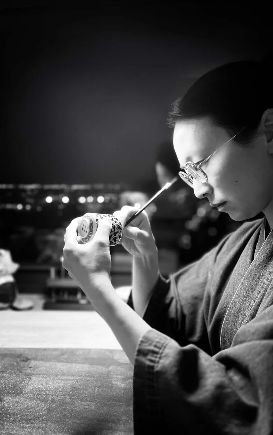 A woman wearing glasses and traditional Japanese clothing, possibly a kimono, is painting on a small ceramic object with a fine brush at night.