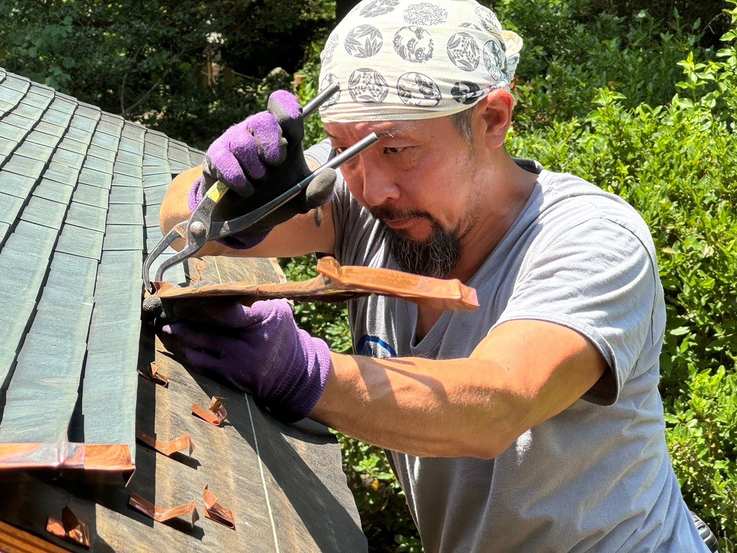 A man wearing a white bandana and purple gloves is using a caulking gun to apply sealant to a roof with green shingles, surrounded by greenery.
