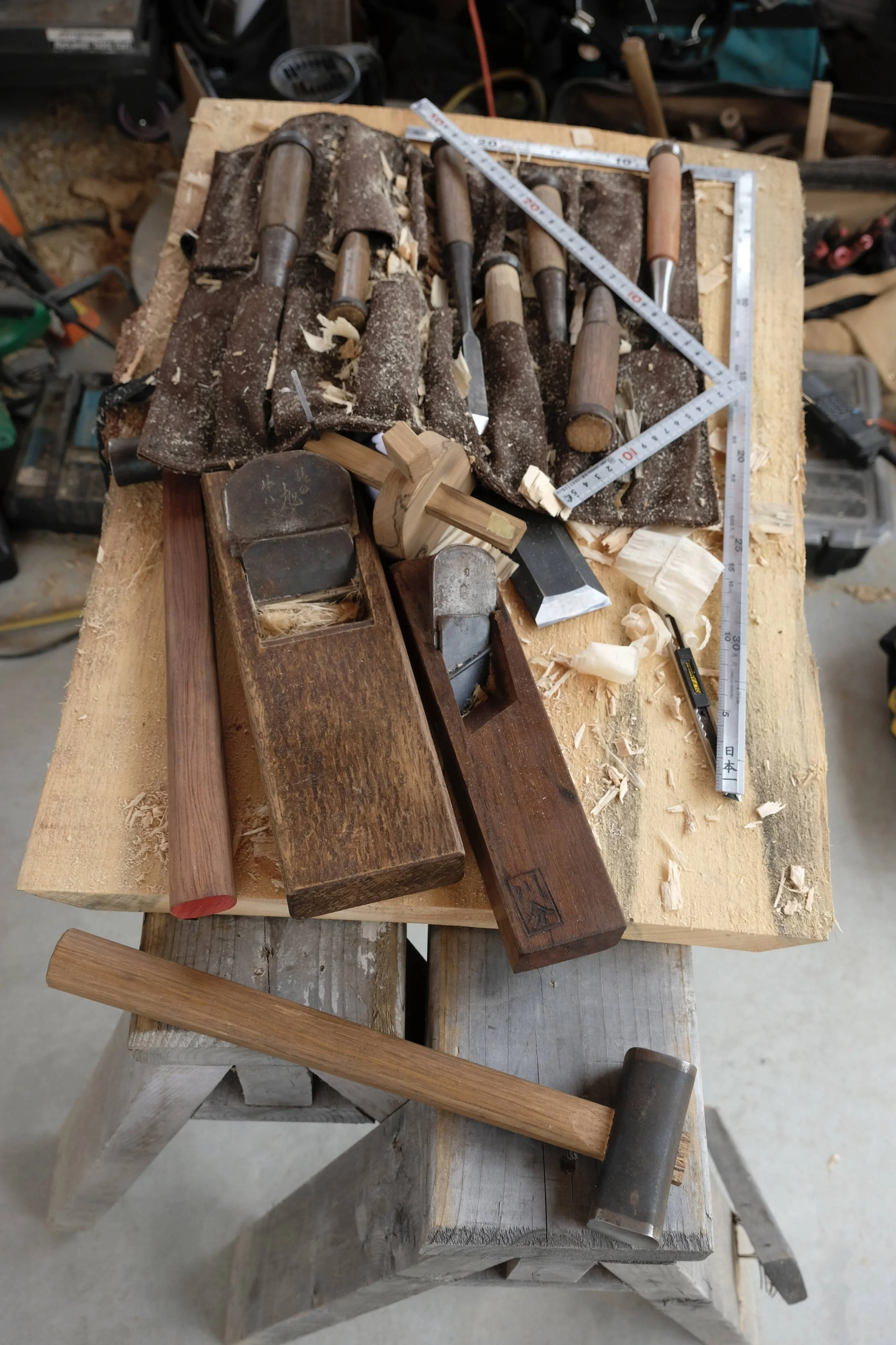 A woodworking workbench with various hand tools including hand planes, chisels, a hammer, a square, and a measuring tape, surrounded by wood shavings and scraps in a woodworking workshop.