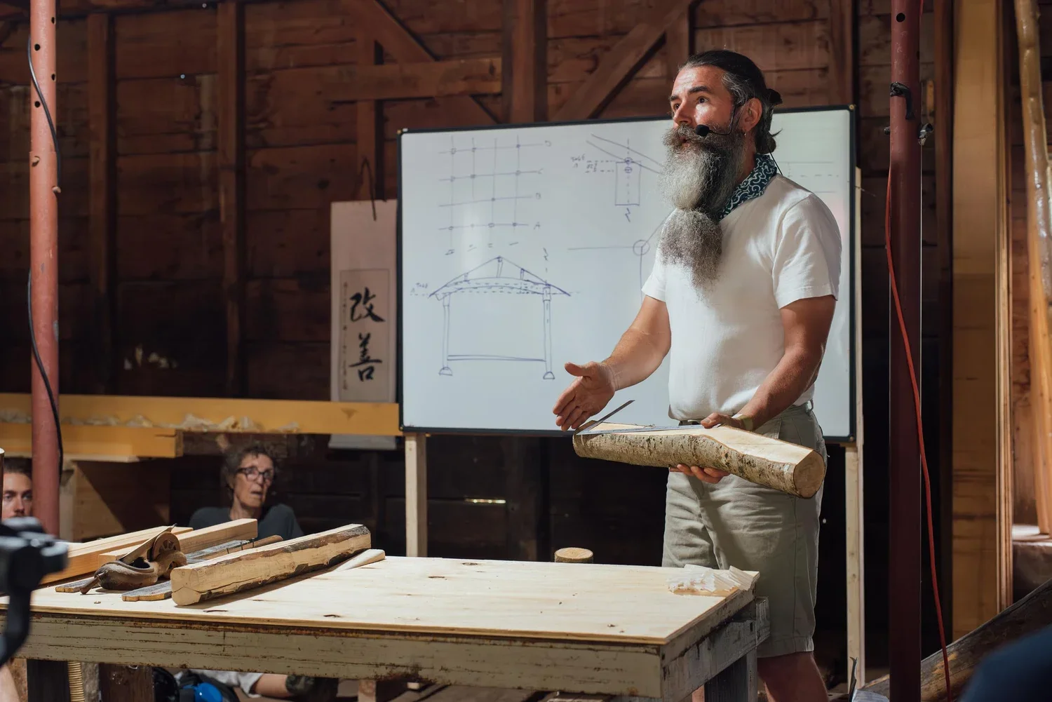 A man with a gray beard, wearing a white t-shirt and shorts, is giving a presentation in a woodworking shop. He is holding a large log and is gesturing with his hands. Behind him, a whiteboard displays sketches of a structure and technical drawings. Several wooden logs and tools are on the table in front of him, and there are attendees sitting and listening.