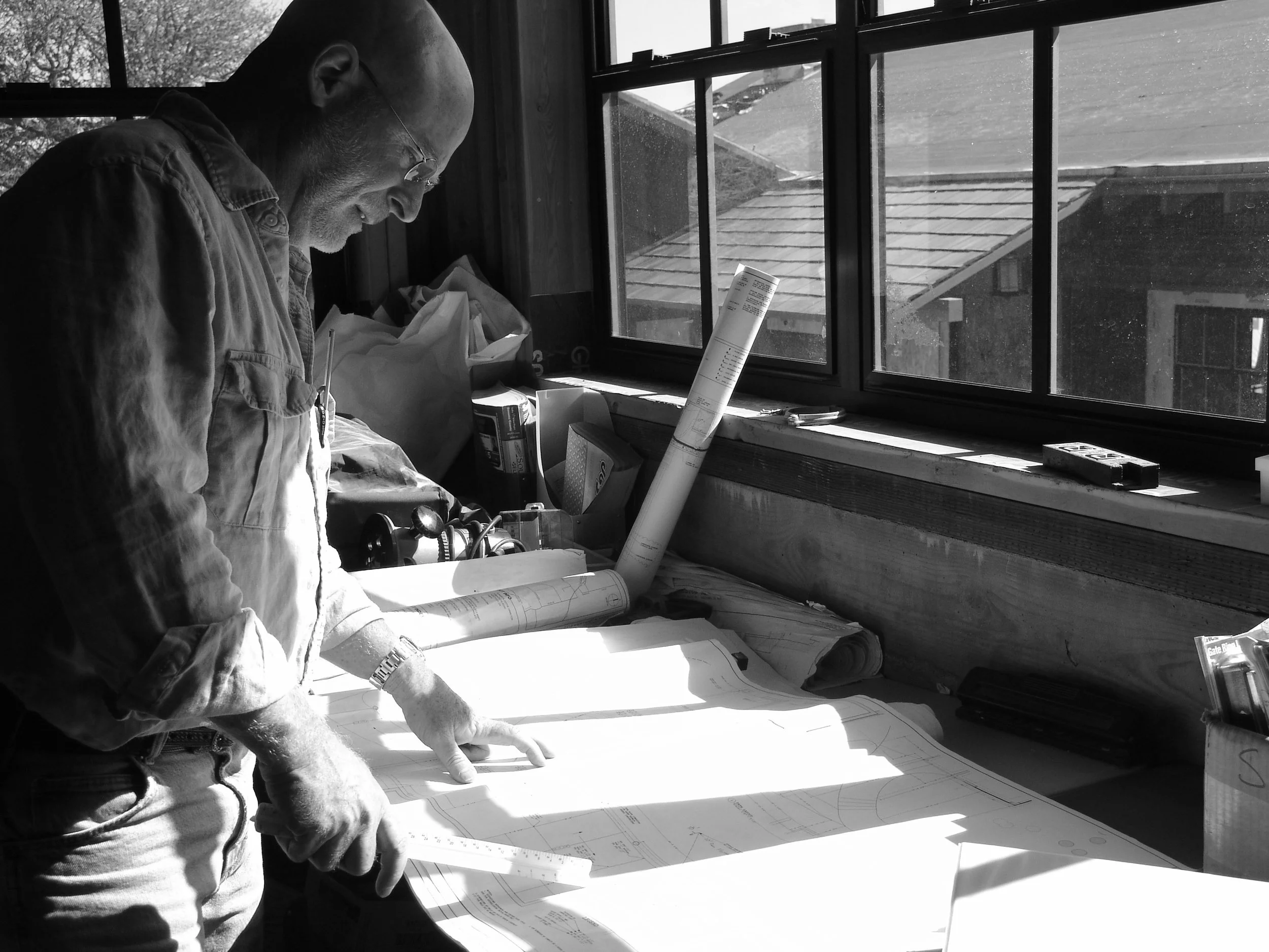 A bald older man with glasses wearing a light blue button-up shirt standing behind a wooden table in a workshop with various wooden items and a machine in the background.