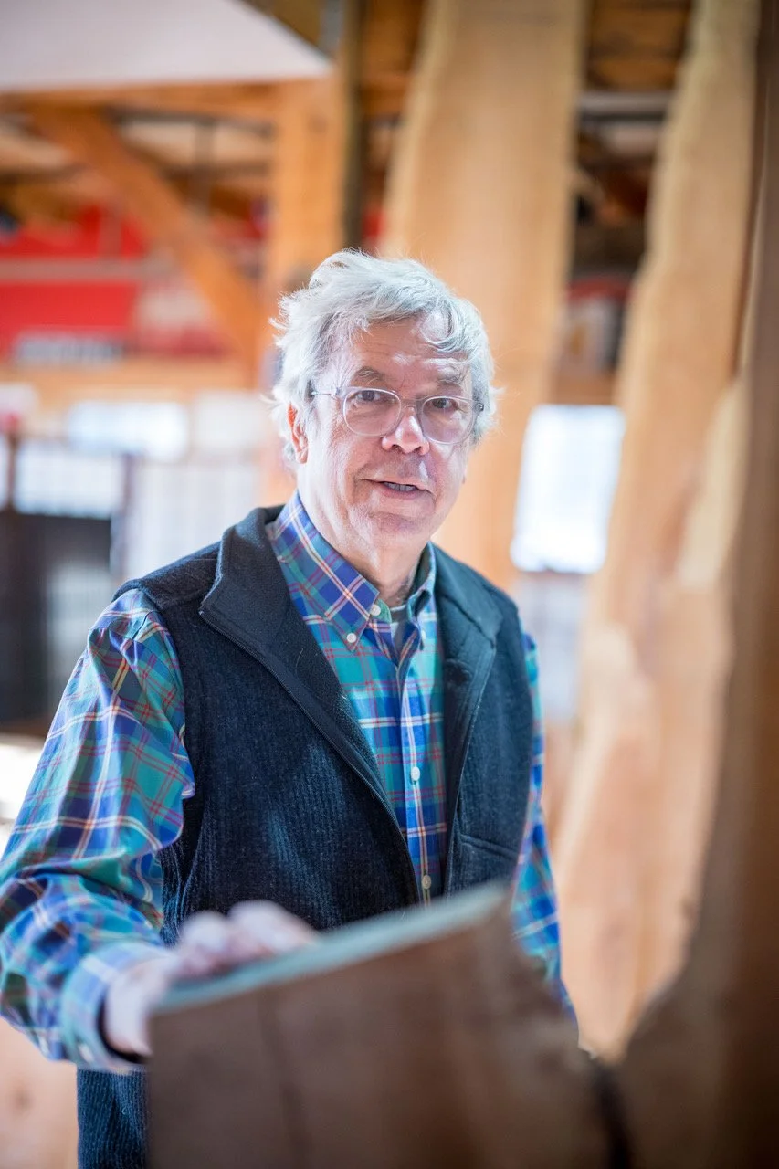 An older man with glasses and gray hair, wearing a plaid shirt and a black vest, is looking at the camera inside a woodworking workshop.