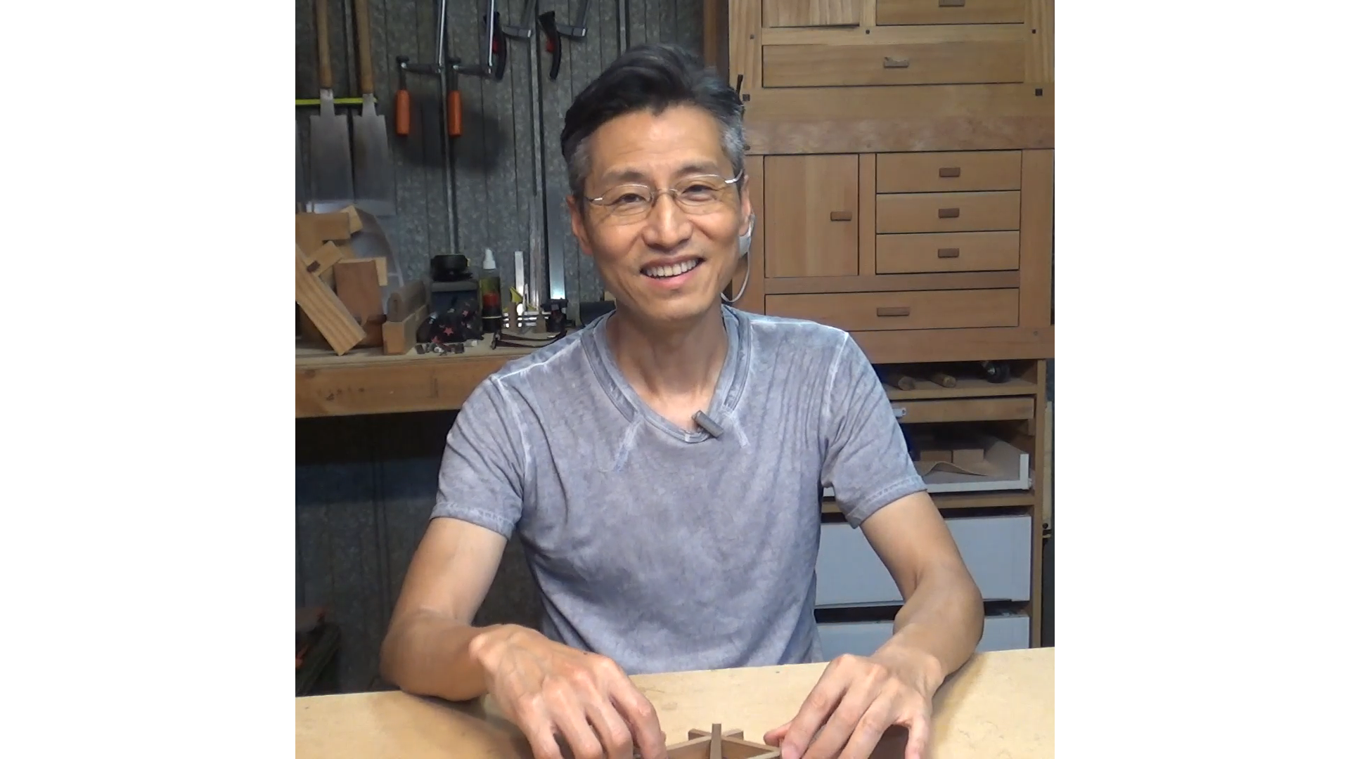 A smiling man with glasses sitting at a woodworking workbench in a workshop, surrounded by tools and wooden furniture.