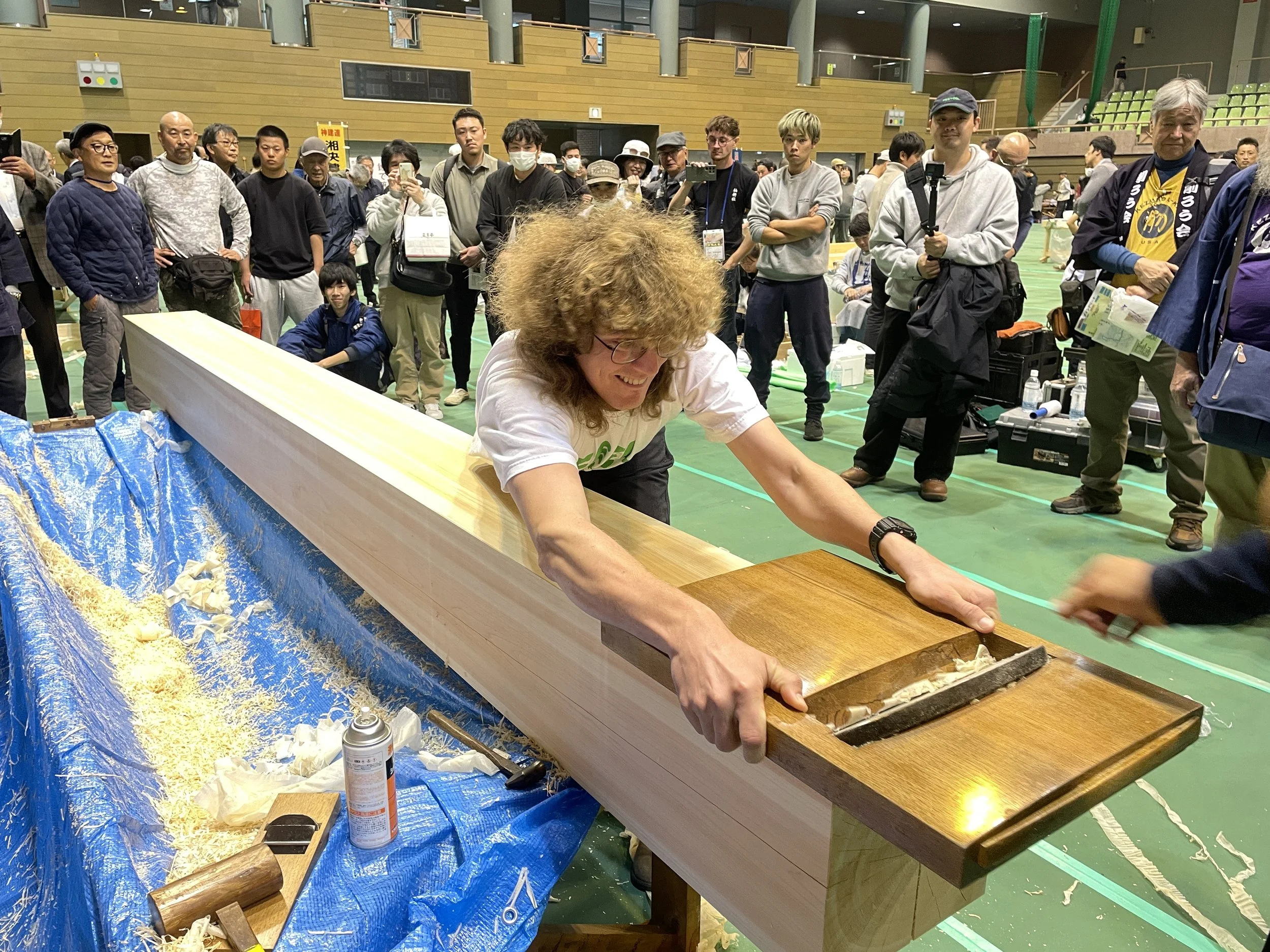 A young man with curly hair, glasses, and a watch is posing outdoors. He's wearing a blue Japanese festival jacket with white symbols and a black shirt underneath. The background shows an outdoor setting with tents, cars, and people.