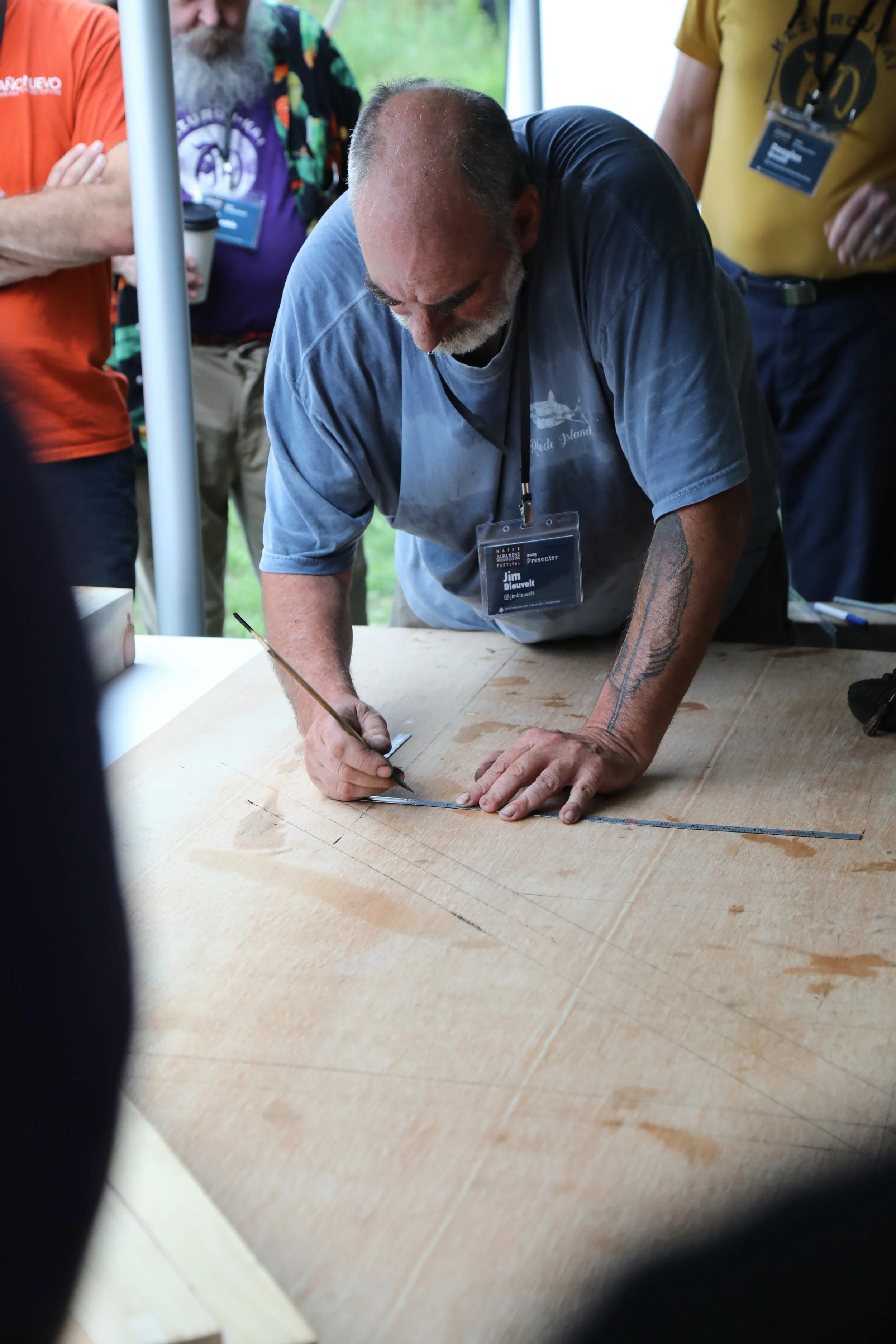 Man marking lines on a piece of wood with a pencil, with several people standing around watching, in an indoor setting.