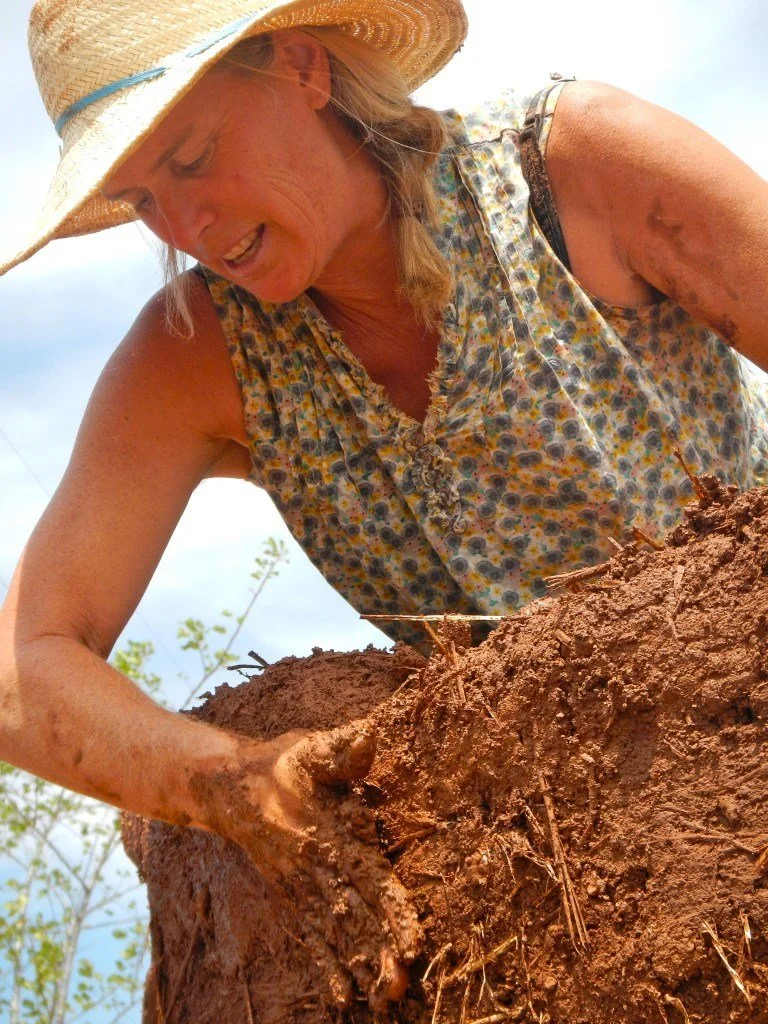 Woman in a floral sleeveless shirt and straw hat digging in the dirt outside.