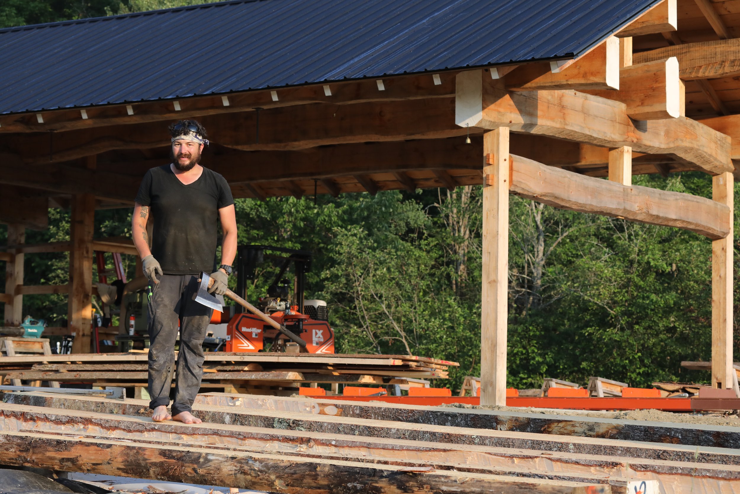 A man stands on lumber at a construction site, holding a hatchet, with a partially built wooden structure in the background and green trees.