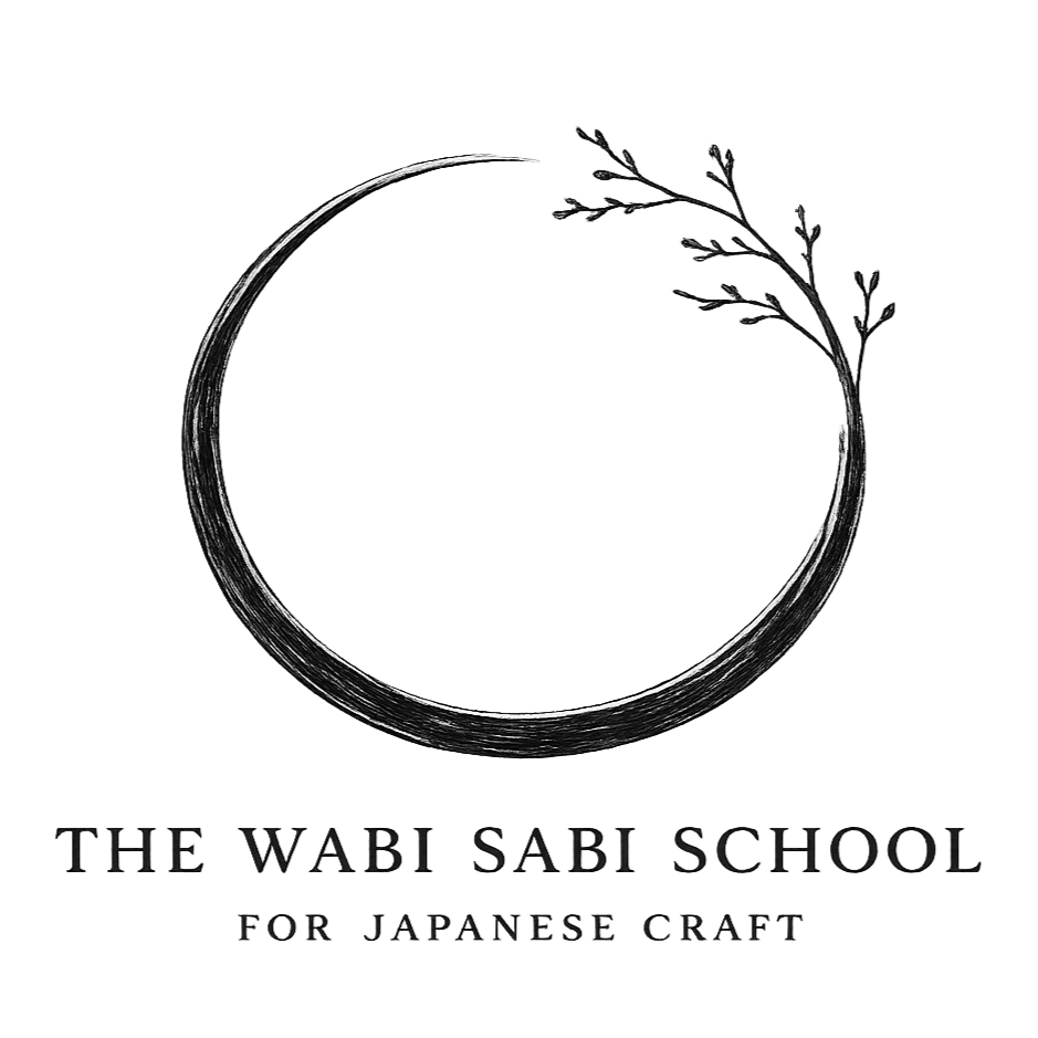 A circular logo with a thin, textured border and a small sprig of leaves extending from the upper right side. Below, the text reads 'The Wabi Sabi School for Japanese Craft' in a minimalist, elegant font.