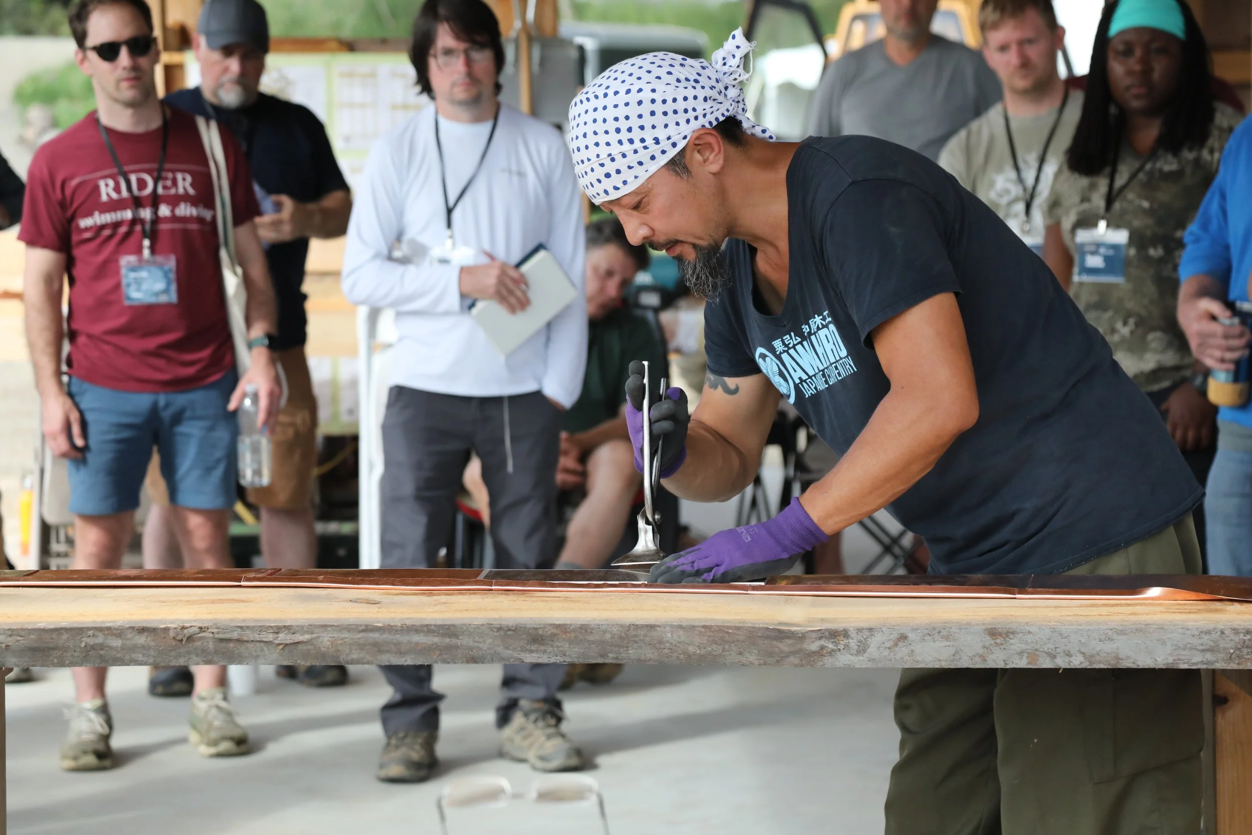 A man wearing a polka dot bandana, black T-shirt, and purple gloves is demonstrating a craft or technique on a wooden surface while a group of people watches attentively in the background.