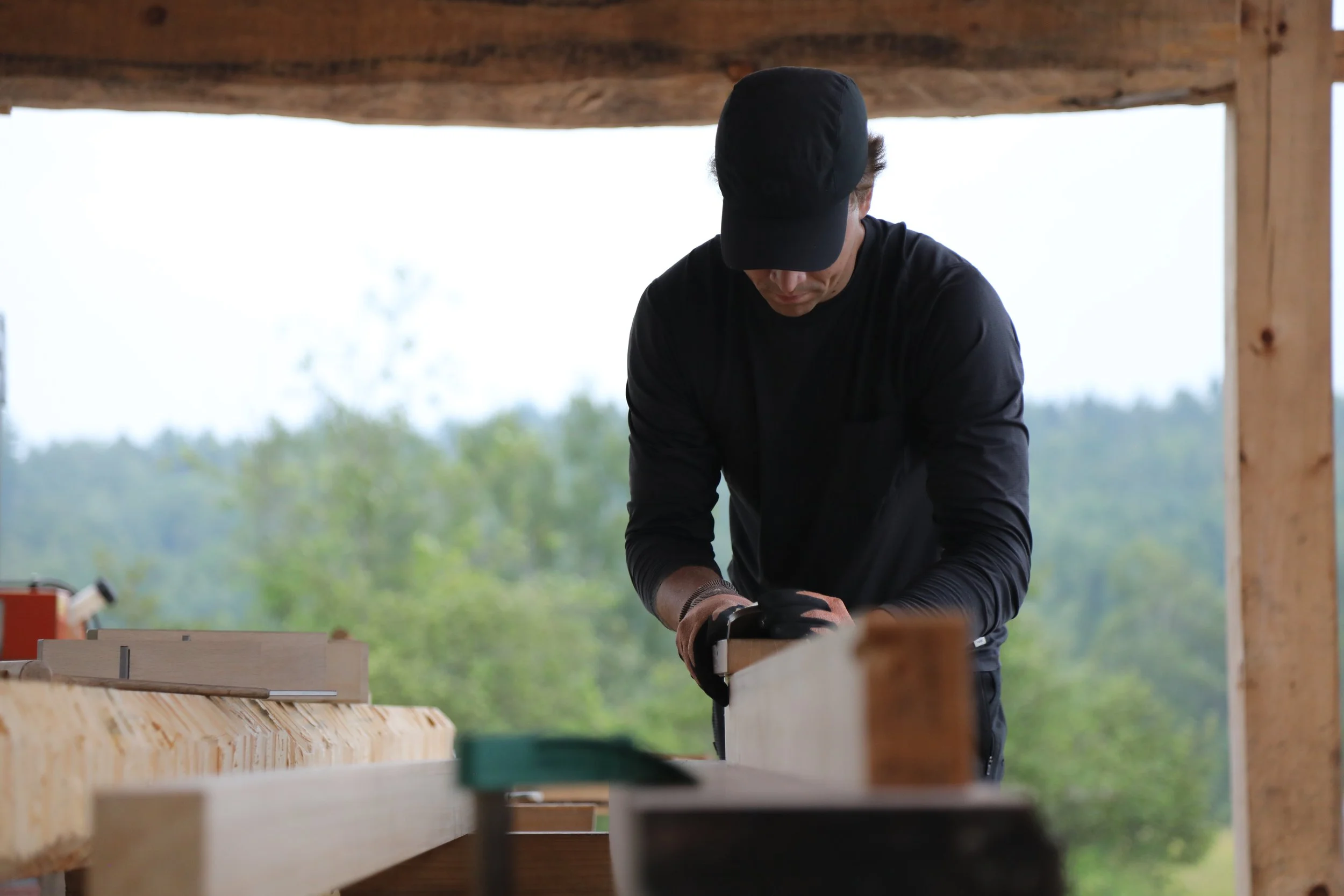 A person wearing a black cap and long-sleeved shirt is working on a woodworking project with a power tool outdoors, surrounded by wooden beams and greenery in the background.