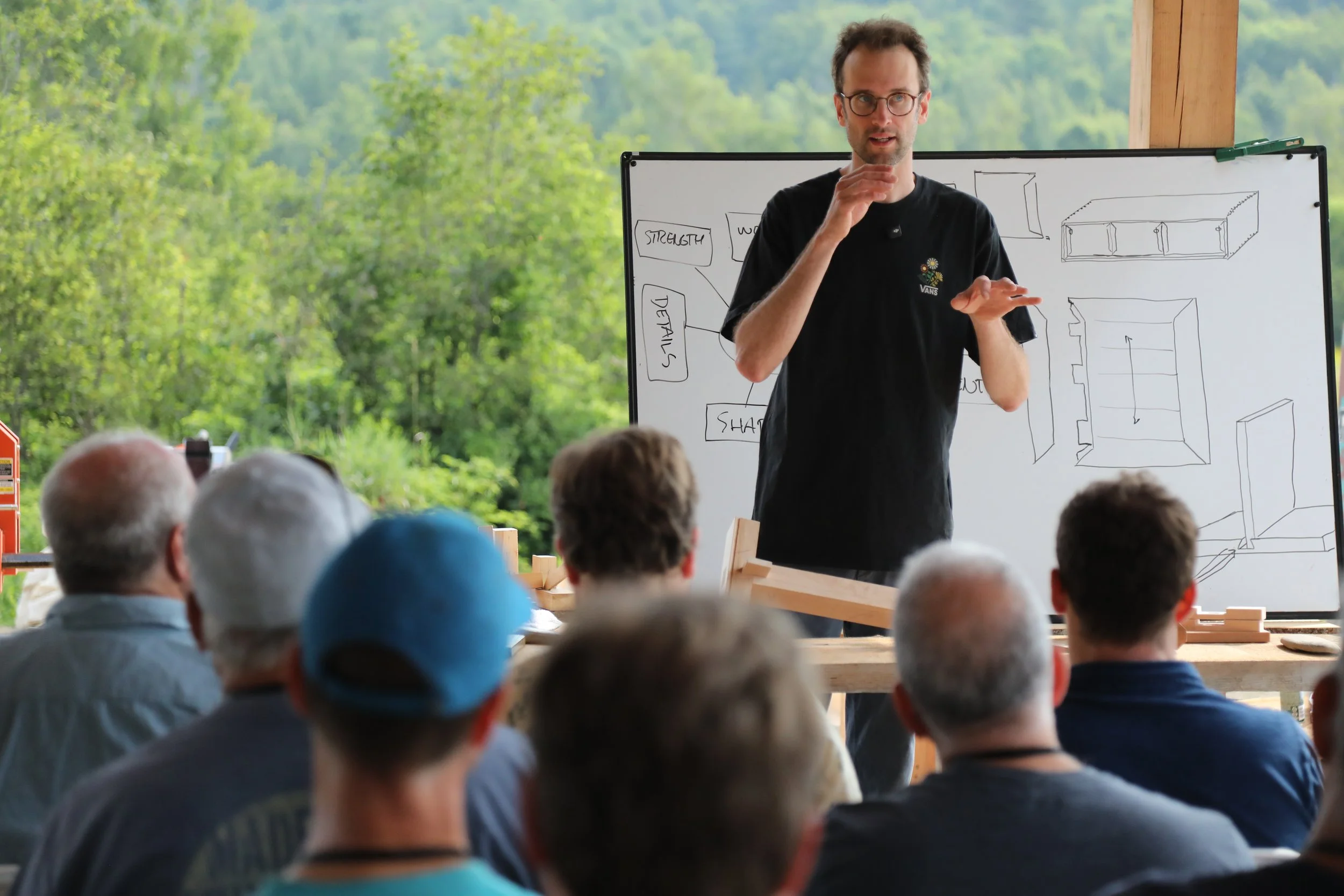 A man giving a presentation to an audience about woodworking or furniture design, with a whiteboard showing sketches and notes, outdoors with green trees in the background.