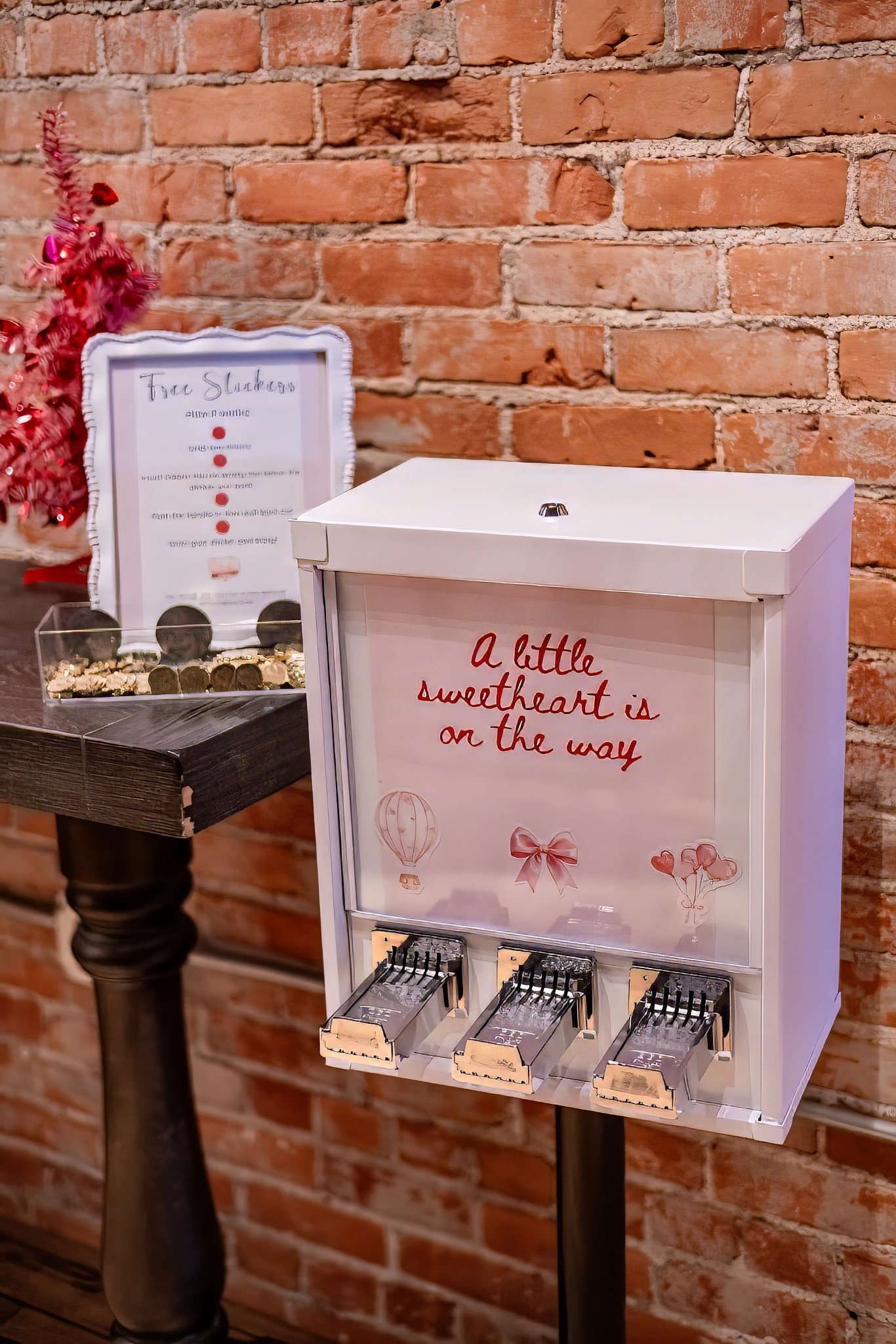 A white box with the message 'a little sweetheart is on the way' decorated with illustrations of balloons, a bow, and hearts, sitting against a brick wall. There are small boxes of chocolates or candies placed in front of the box.