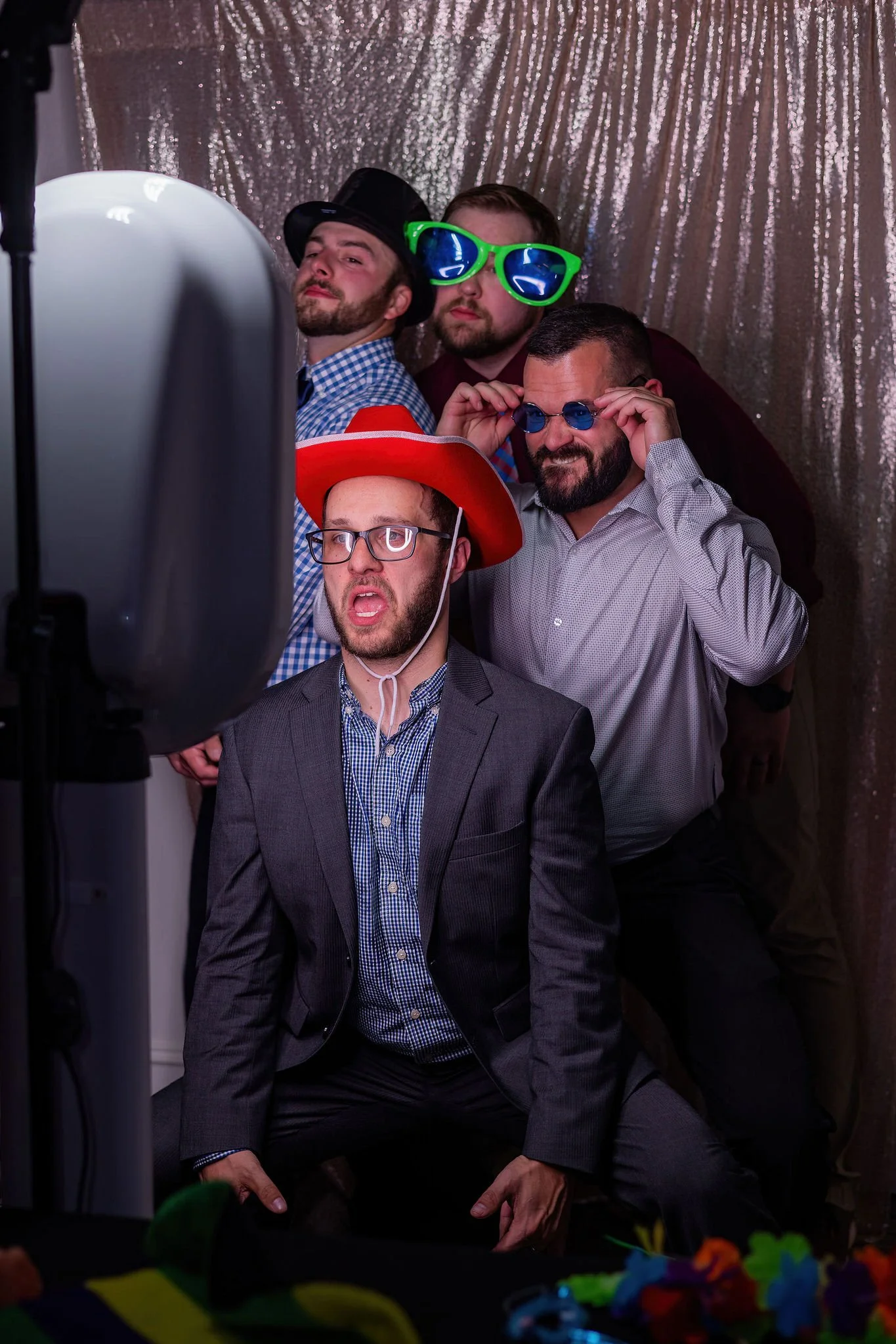 Group of five men dressing in formal and fun accessories taking a photo in a photo booth with a silver sequin backdrop.