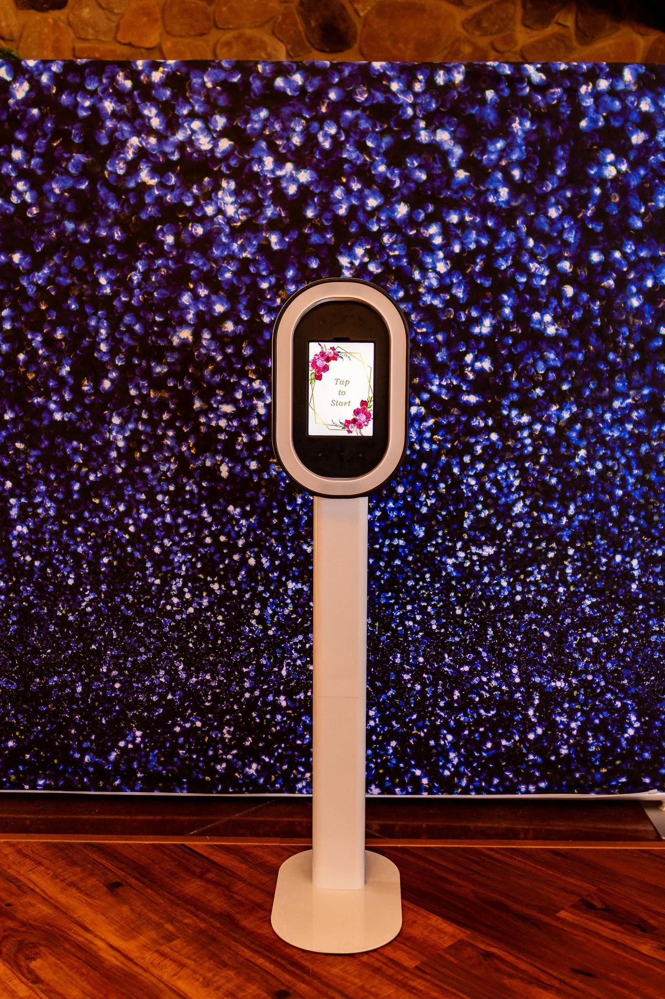 A digital kiosk with a small screen that says 'Tap to Start' is positioned in front of a large floral display in shades of purple and blue, with a brick wall above. The stand is white and placed on a wooden floor.