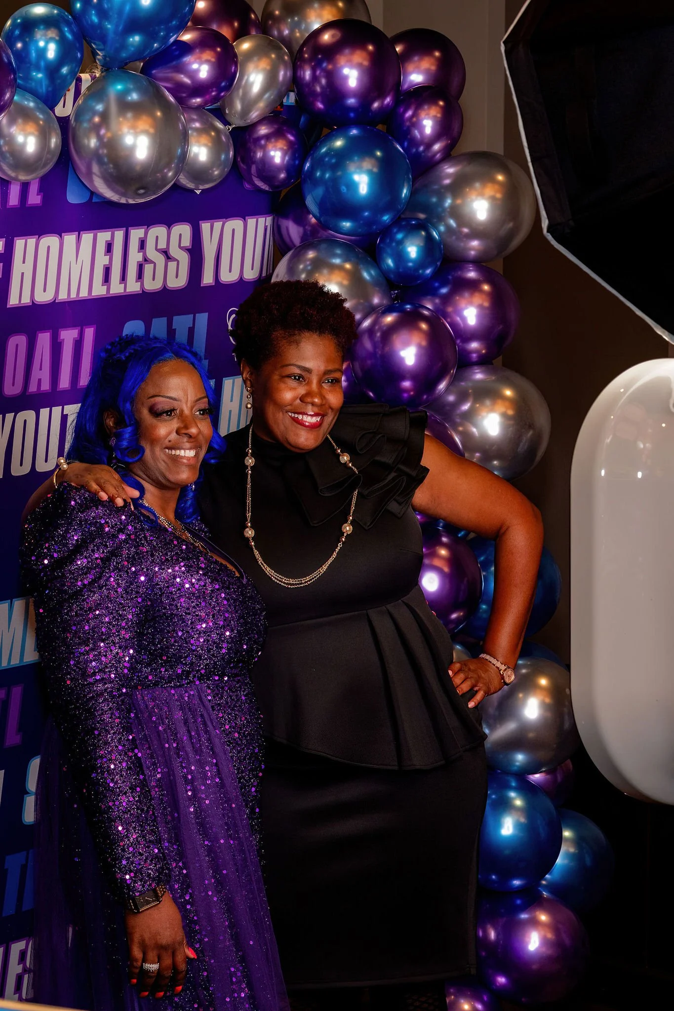 Two women smiling and posing in front of a colorful backdrop with purple, blue, and silver balloons. One woman is wearing a purple sequined dress, and the other is in a black dress with ruffled sleeves, surrounded by balloons.
