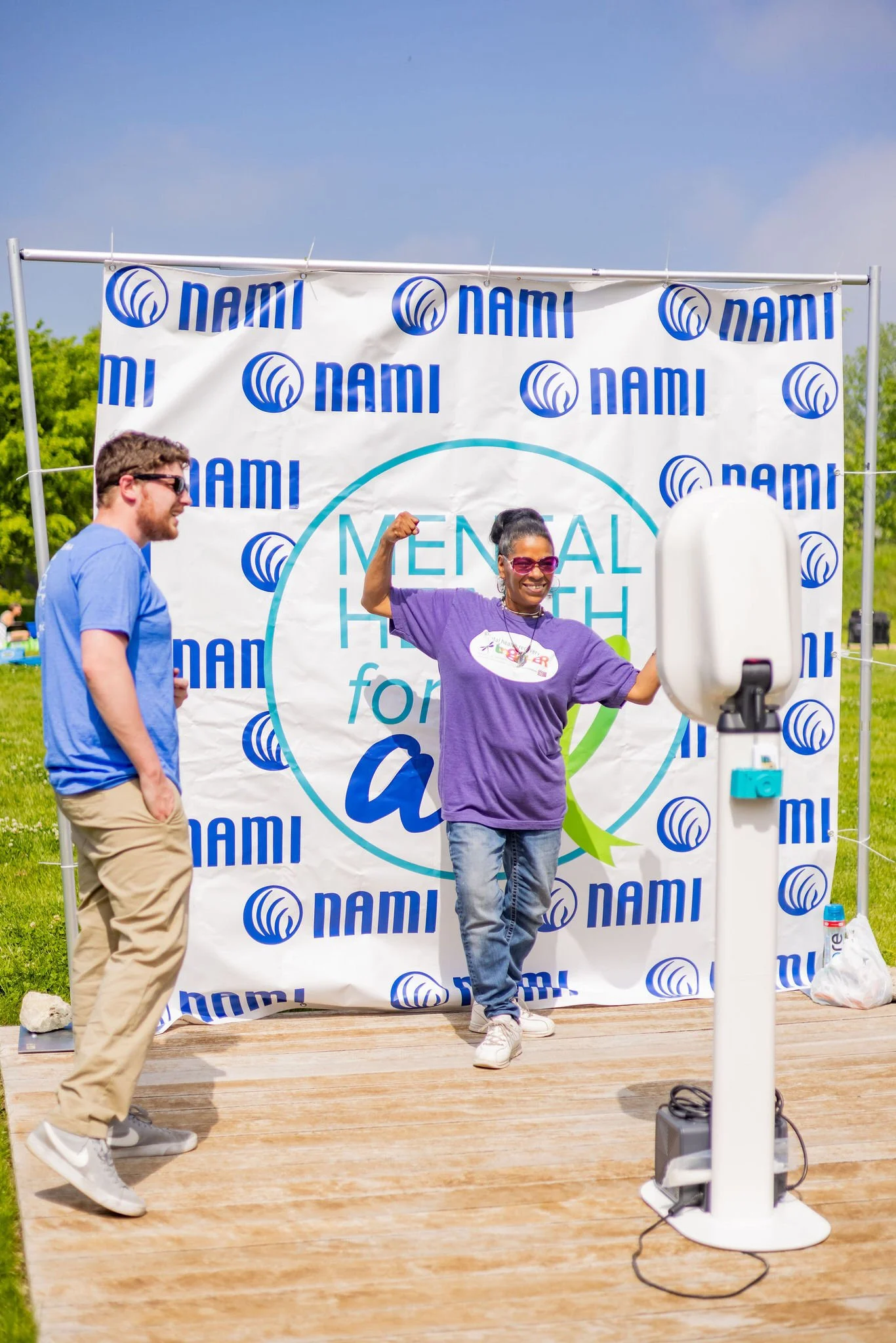 A woman flexing her arm and smiling on stage at an outdoor event, with a man standing nearby, in front of a backdrop with the NAMI logo and the text 'Mental Health for All'.
