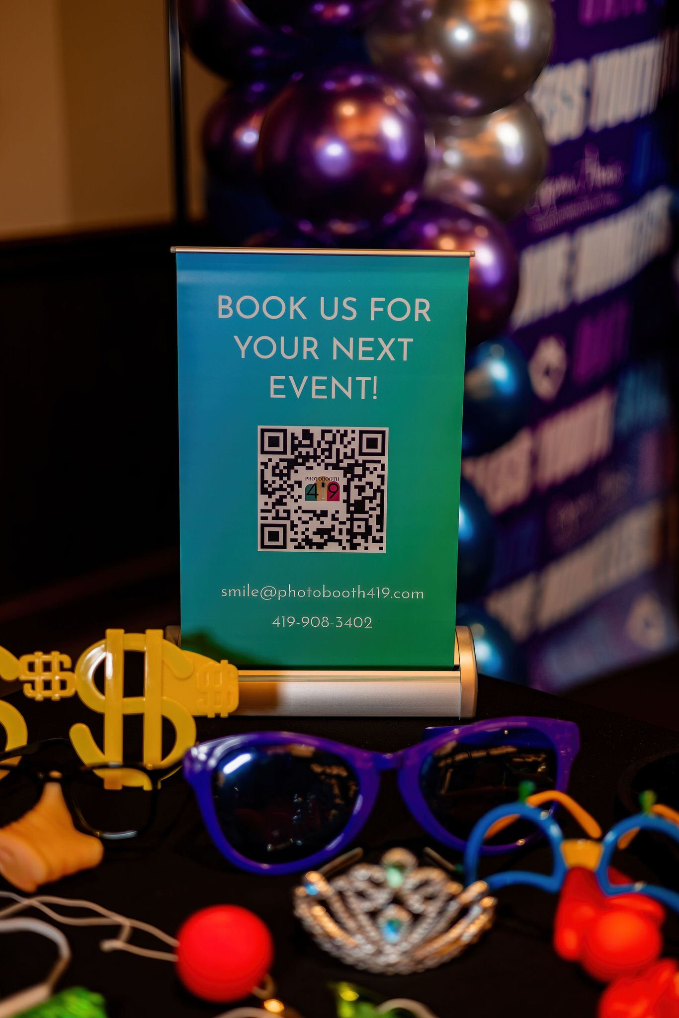A sign offering to book events via a QR code with contact information, surrounded by colorful sunglasses, decorative jewelry, and party props on a table.