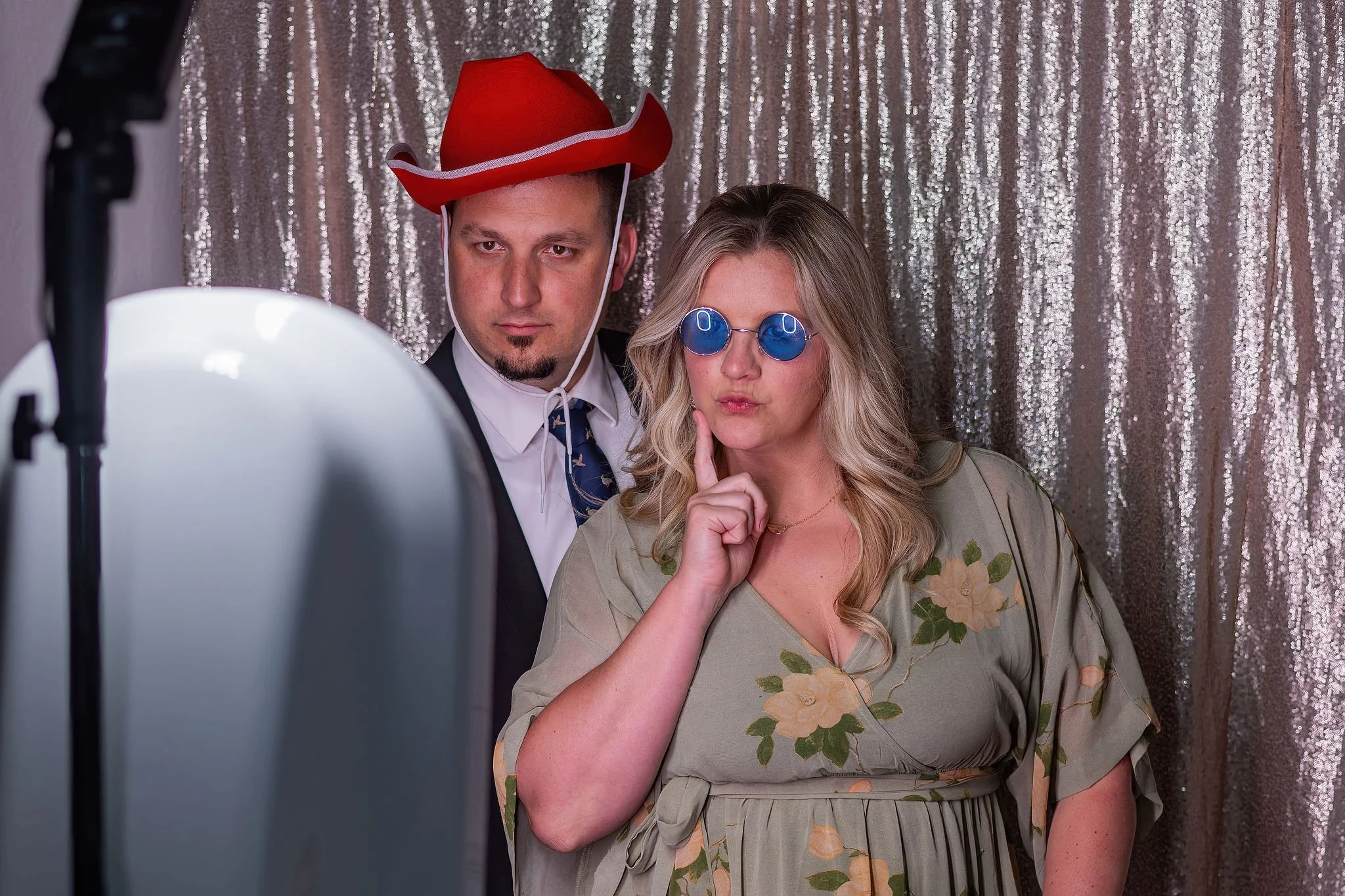A man and a woman posing for a photo booth picture, with a sparkly silver backdrop. The man is wearing a red cowboy hat and a suit with a tie, and has a serious expression. The woman is wearing round blue sunglasses, a floral dress, and is holding he