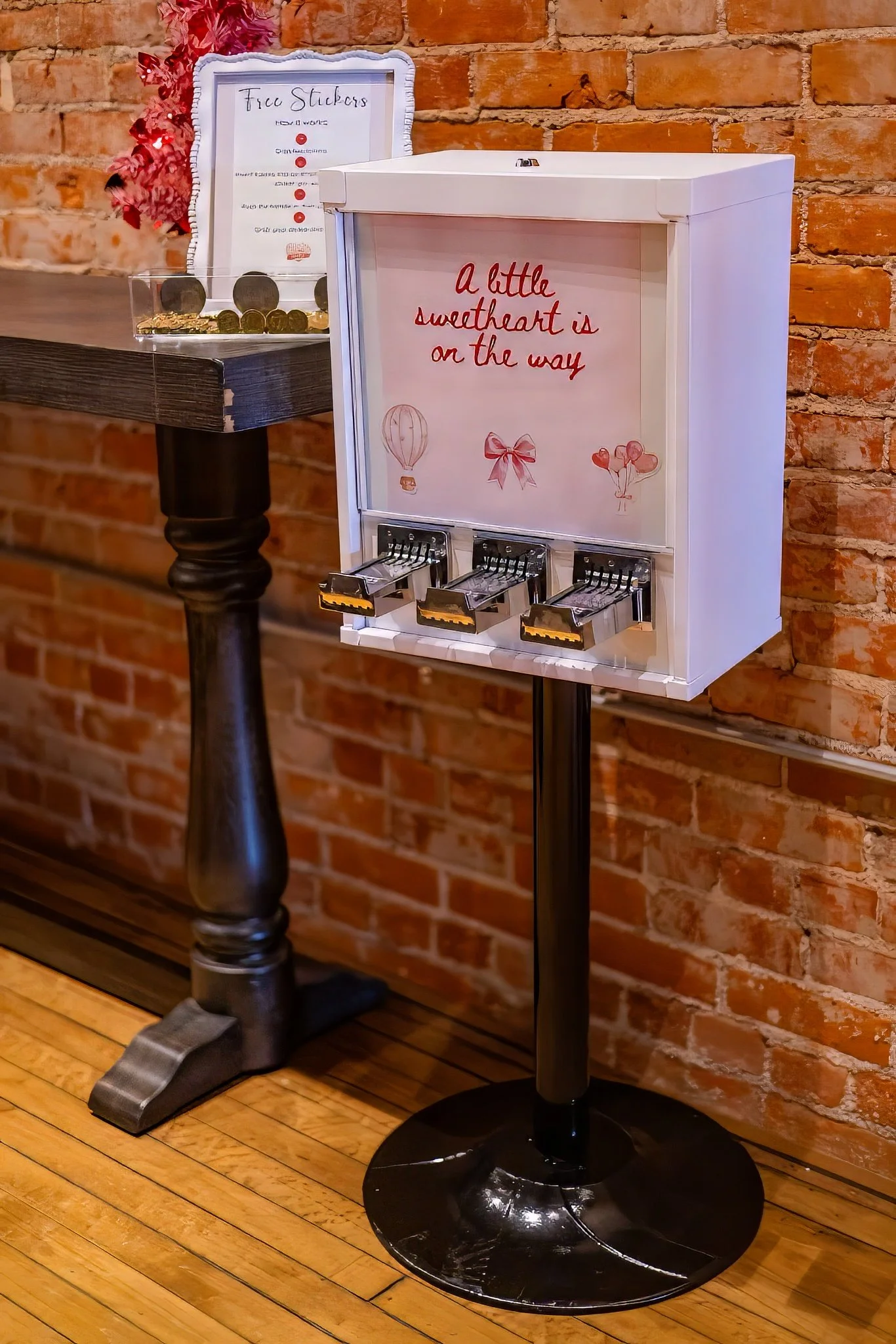A white candy dispenser with a sign that says, 'a little sweetheart is on the way,' featuring images of a hot air balloon, a bow, and heart balloons. The dispenser is placed on a black stand against an exposed brick wall.