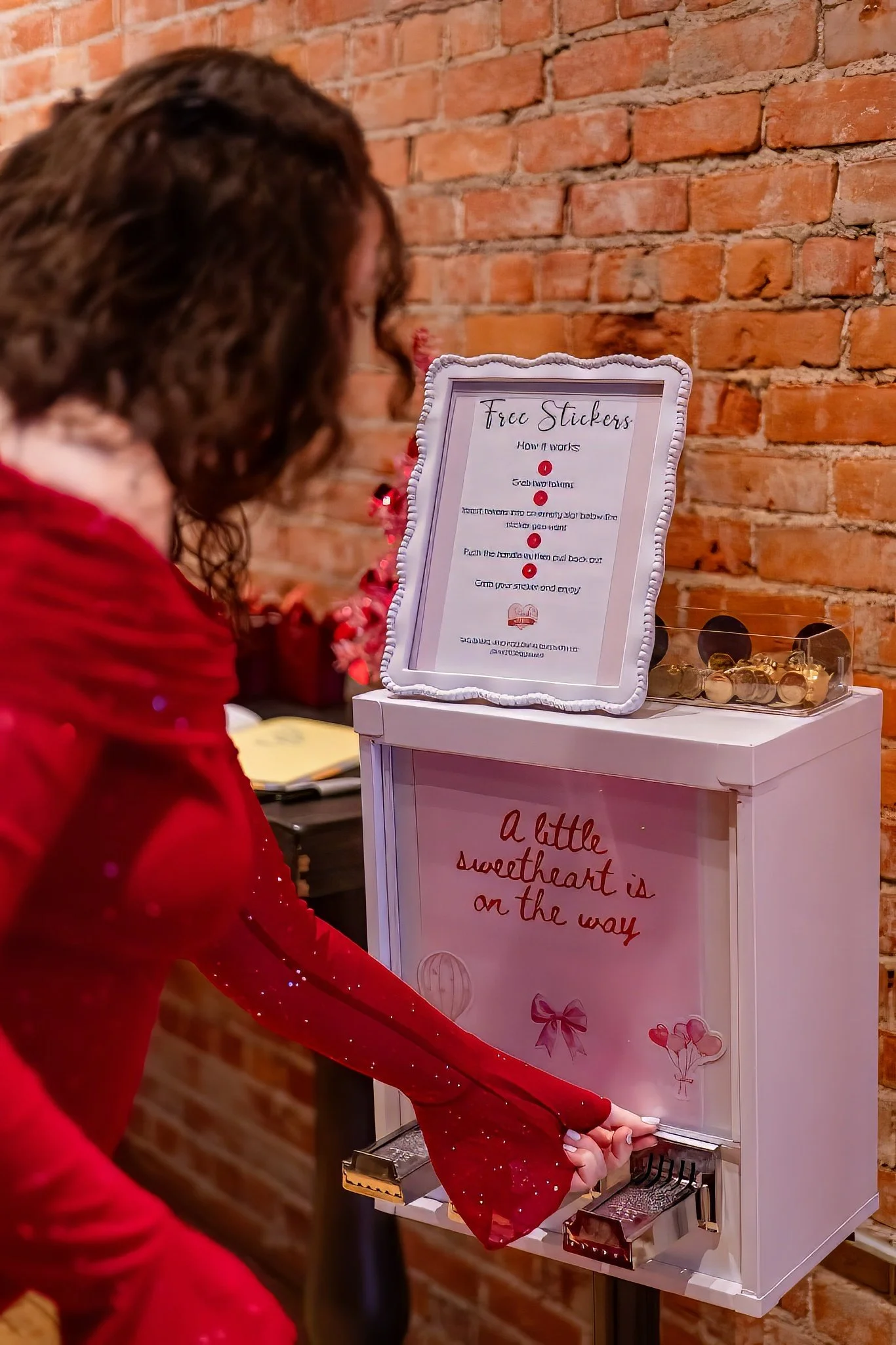 A woman in a red dress is using a coin-operated photo booth that has a decorated back with pink and red accents, including a sign that says "A little sweetheart is on the way."