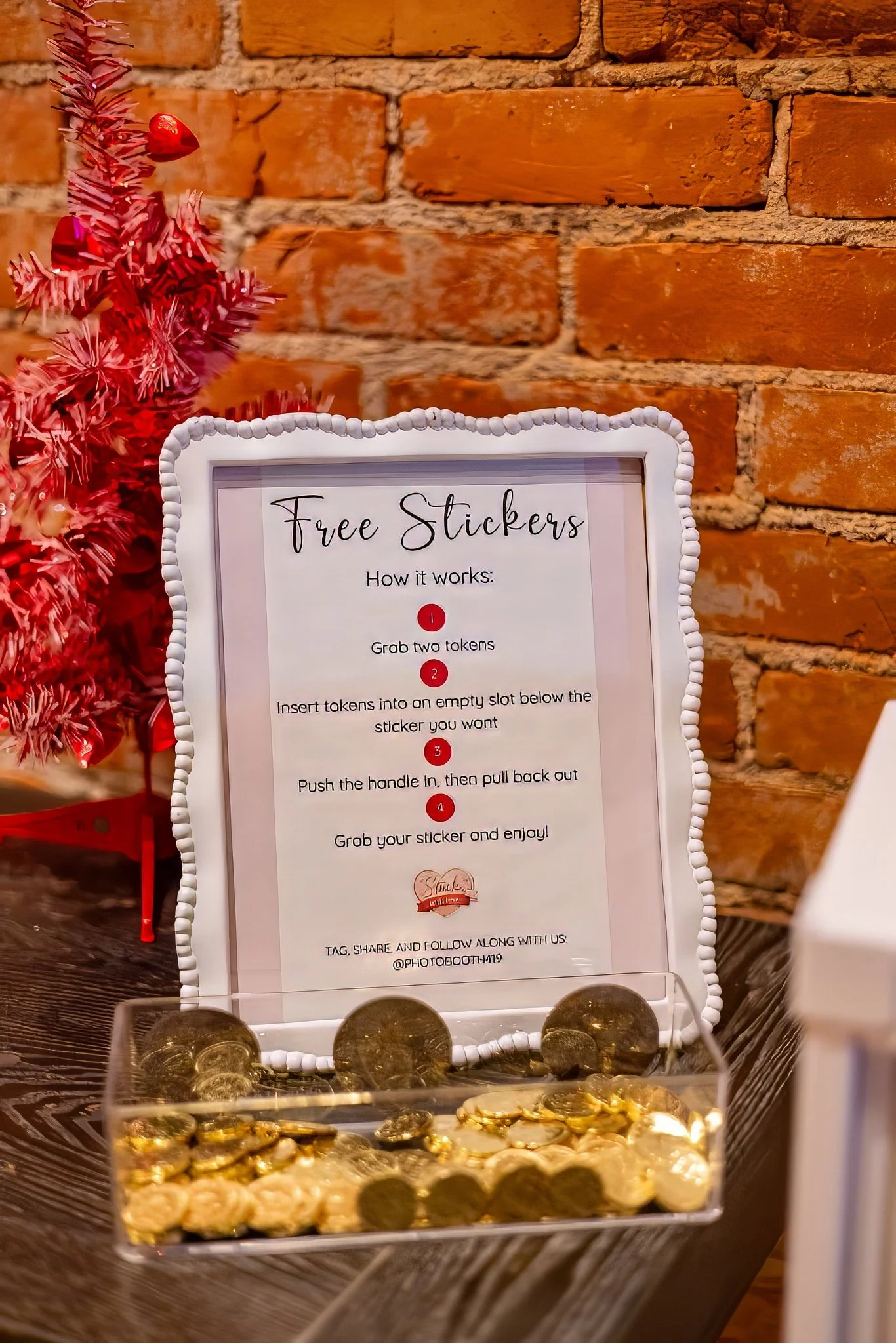 Sign with instructions for free stickers next to a pink mini Christmas tree, with a container of gold and silver coins in front. The sign explains how to take a sticker using tokens.