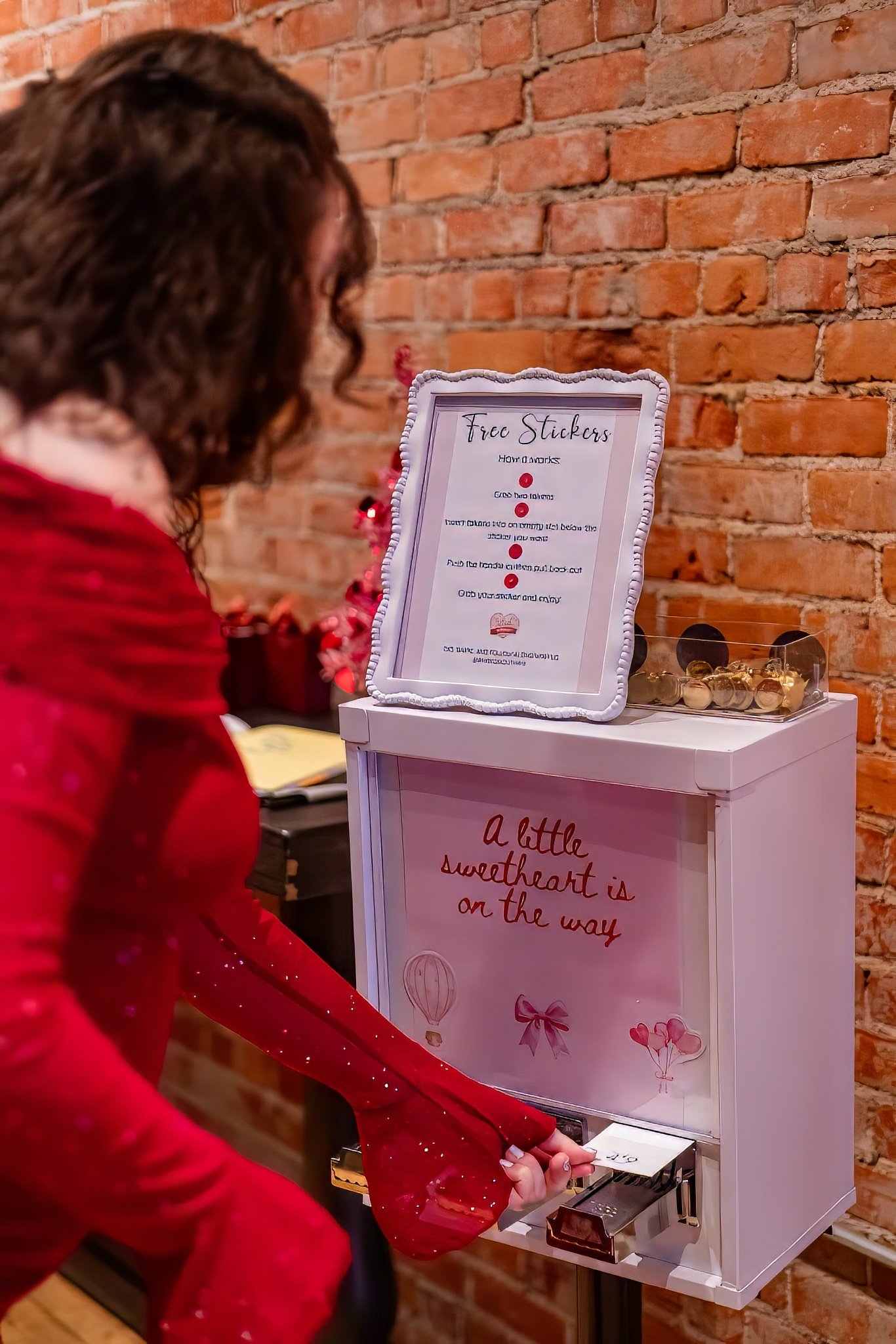 A woman in a red dress is reaching into a white box labeled 'a little sweetheart is on the way' to drop in a card or note. The box has pink and red decorations, including balloons and ribbons. On top of the box is a sign offering free stickers with i