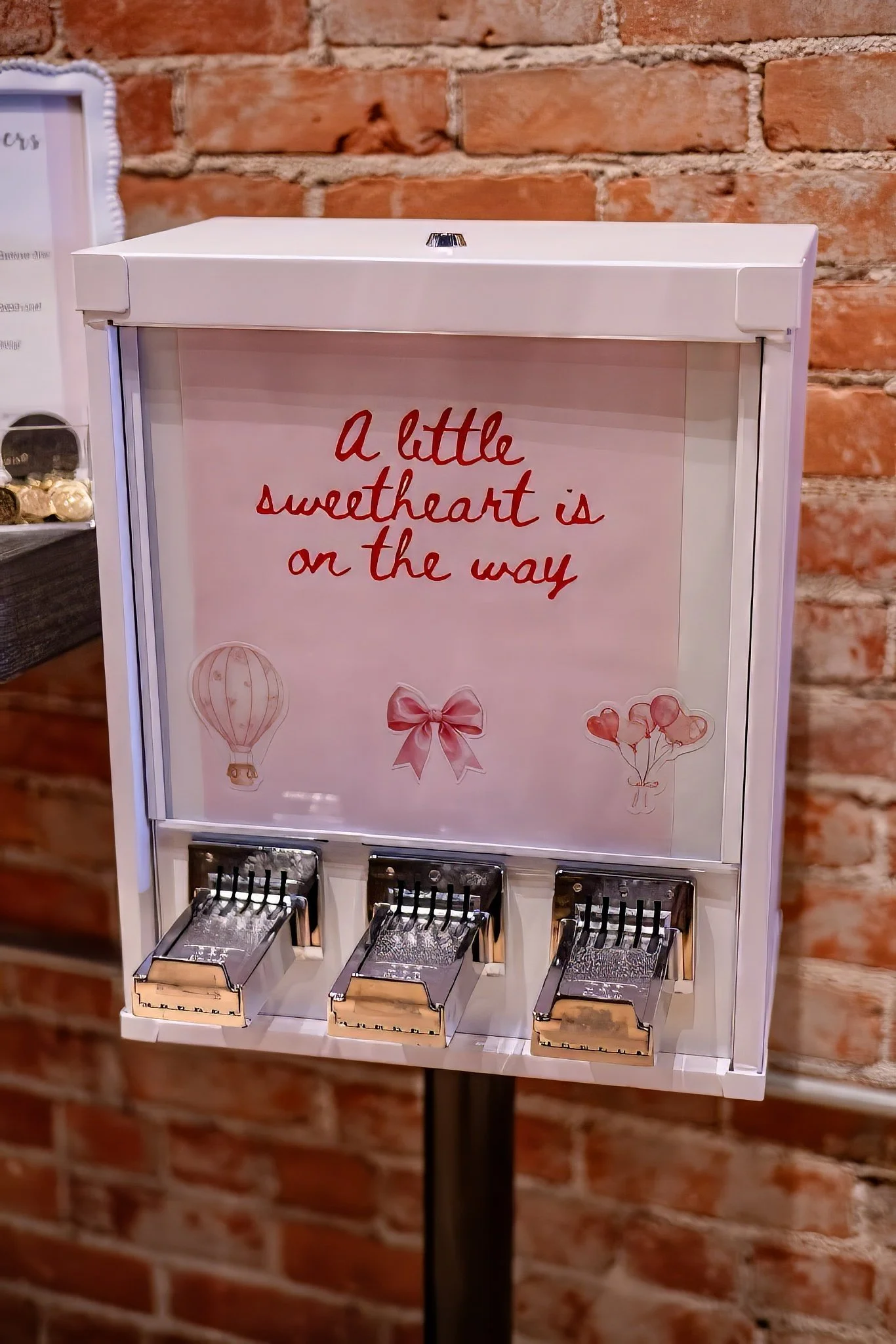 A white candy dispenser with a sign that reads 'A little sweetheart is on the way,' decorated with pink illustrations of a hot air balloon, a bow, and heart-shaped balloons, placed against a brick wall.