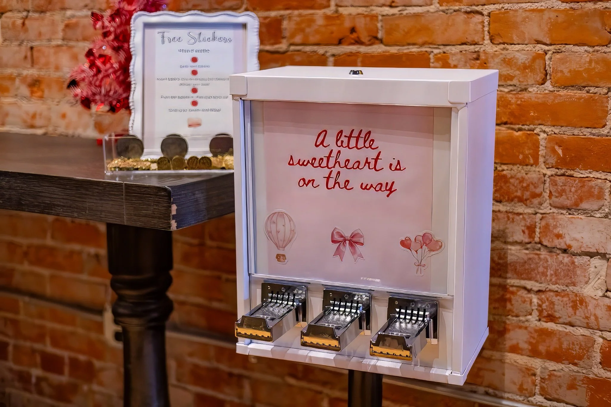 A pink message board with the text 'a little sweetheart is on the way' and cute Valentines Day-themed illustrations, including a hot air balloon, a bow, and heart balloons, positioned next to a wooden table with a red decorative item and a glass container filled with chocolates, against a brick wall background.