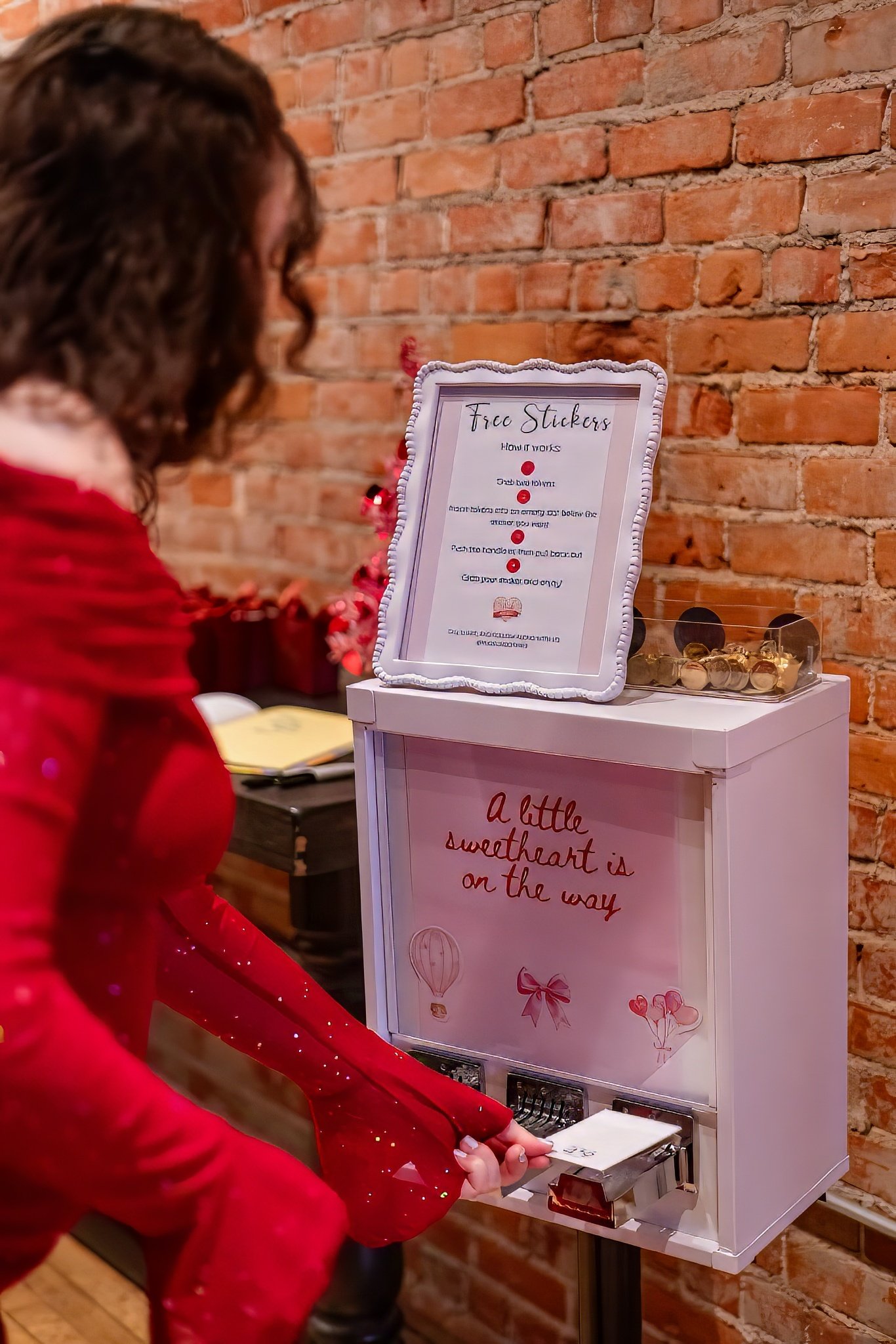 A woman in a red dress adds money to a donation box at a Valentine's themed event, with a brick wall in the background.