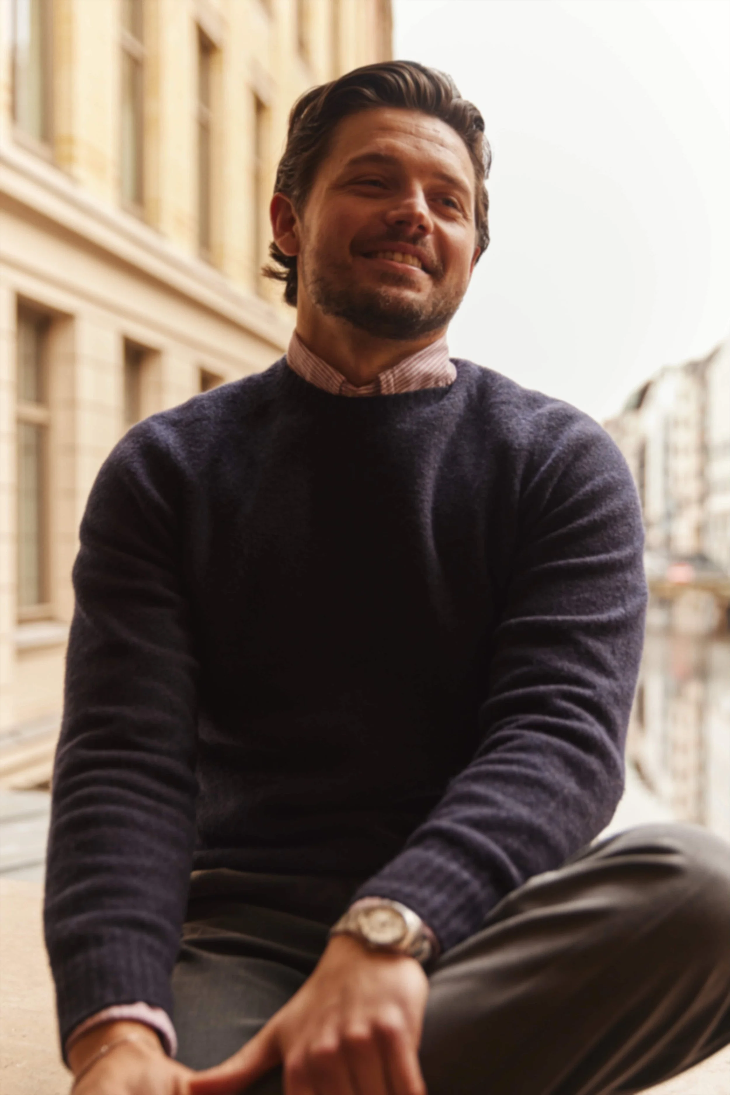 Ein lächelnder Mann sitzt auf einer Stadtstraße mit alten Gebäuden im Hintergrund, trägt einen dunkelblauen Pullover und ein Armbanduhr.
