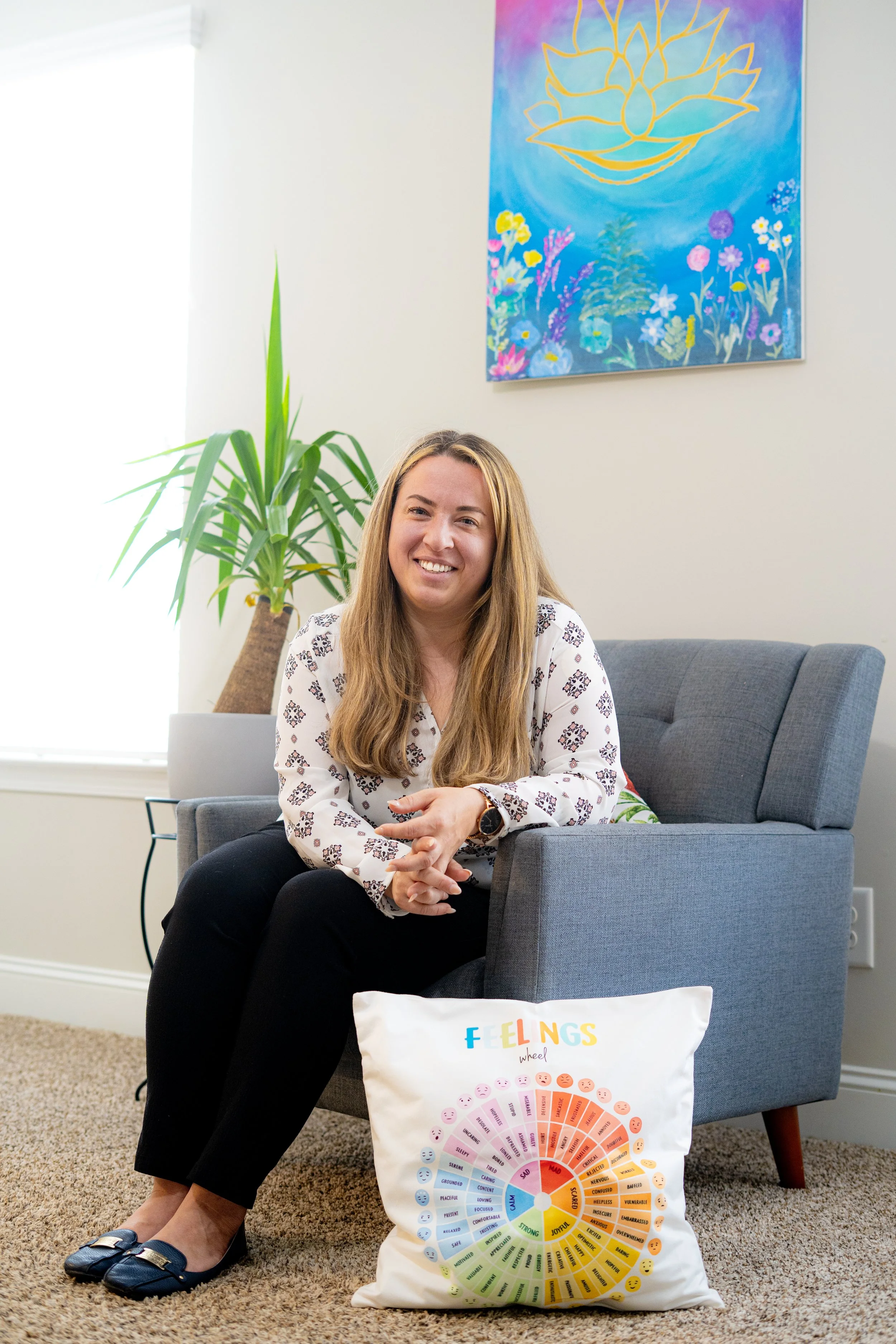 Therapist is sitting on a gray armchair smiling, with a white pillow on the floor in front of her titled 'FEELINGS wheel' showing different emotions. Behind her is a potted plant and a colorful painting of a lotus flower and flowers on the wall.