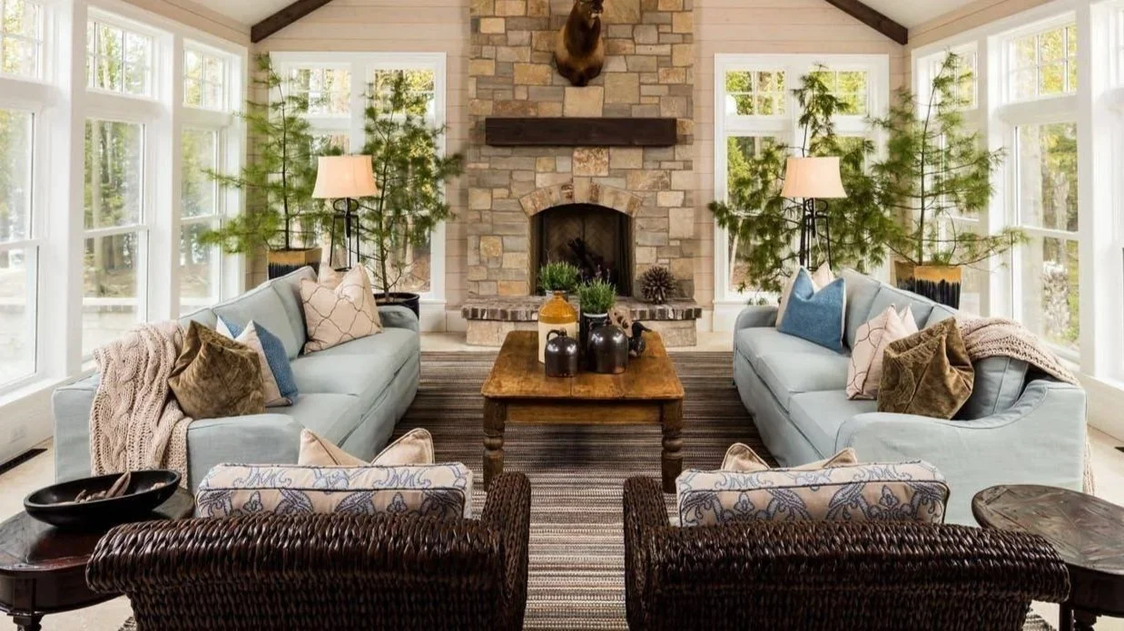 Living room with large windows, two light blue sofas, patterned pillows, a wooden coffee table, a brick fireplace with a mounted animal head, and tall plants near the windows.
