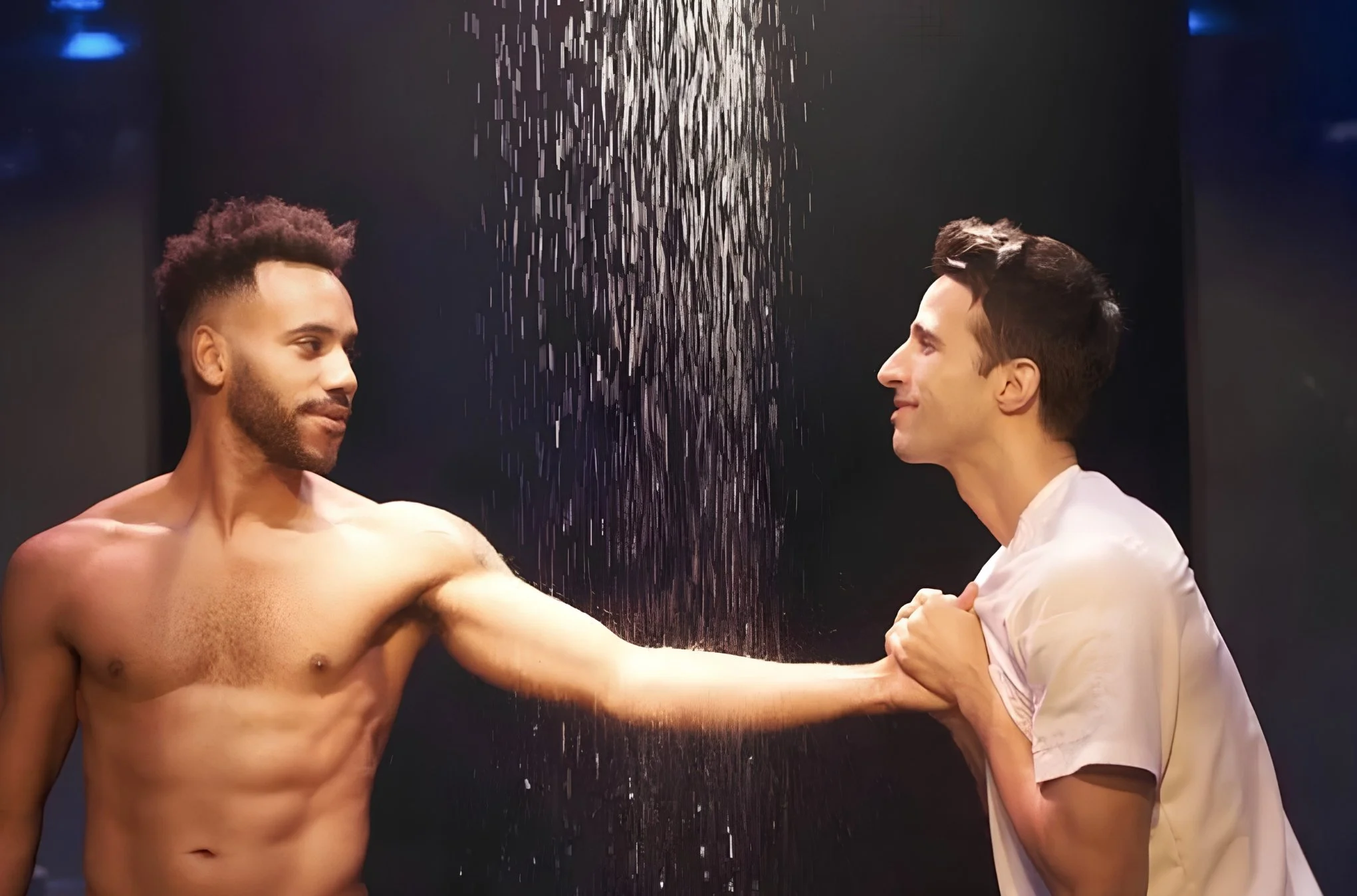 afterglow the play. two men, one shirtless with muscular build and short curly hair, the other wearing a white shirt with short dark hair, facing each other with serious expressions in front of a black background with water falling from the ceiling.
