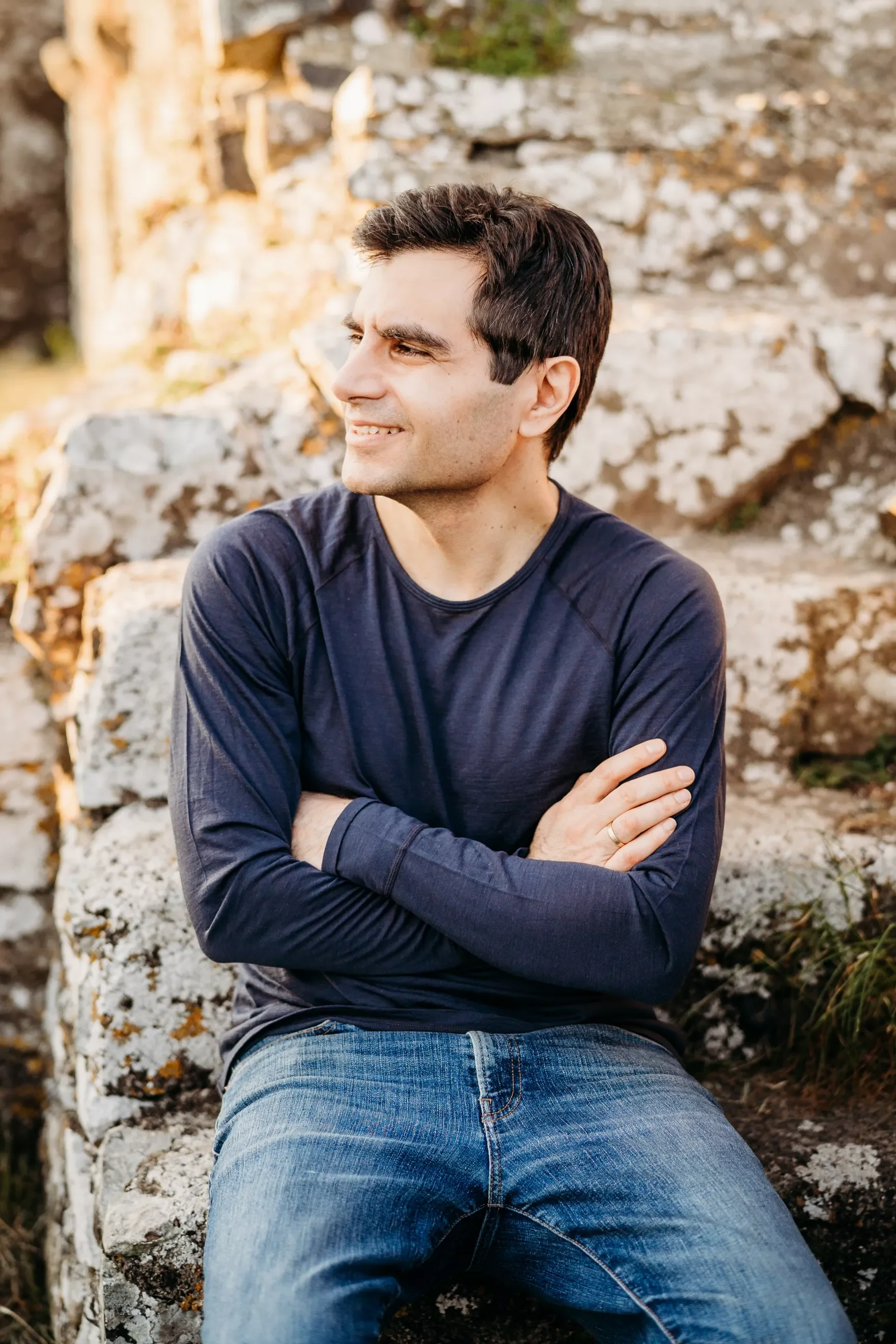 A man sitting on rocks outdoors, wearing a long-sleeve dark shirt and jeans, with arms crossed, looking to the side with a slight smile.