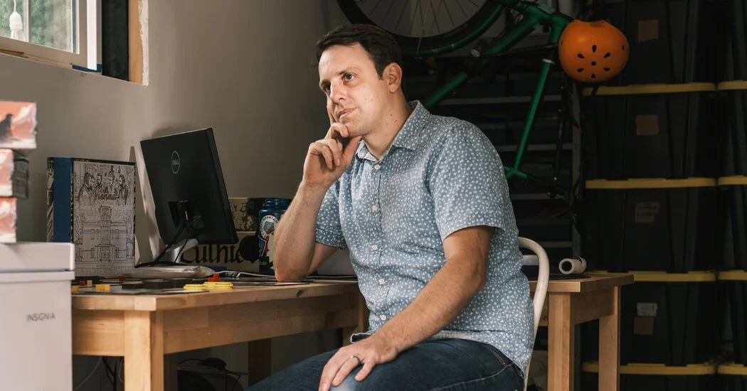 A man sitting at a desk with a thoughtful expression, touching his chin, in a workspace with a computer, shelves, and a green bicycle in the background.