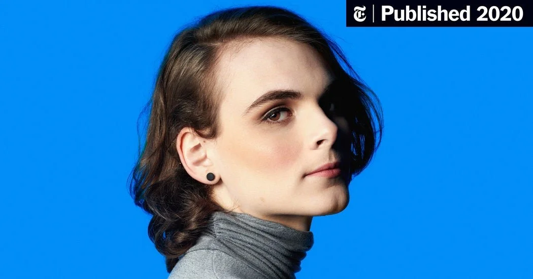 Close-up portrait of a woman with short brown hair, wearing a gray turtleneck and black earrings, against a blue background with a publication mark in the top right corner.
