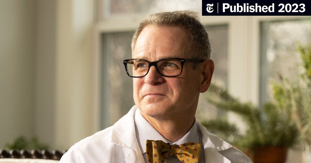 A man wearing glasses, a white lab coat, and a yellow bow tie looking out a window with a thoughtful expression.