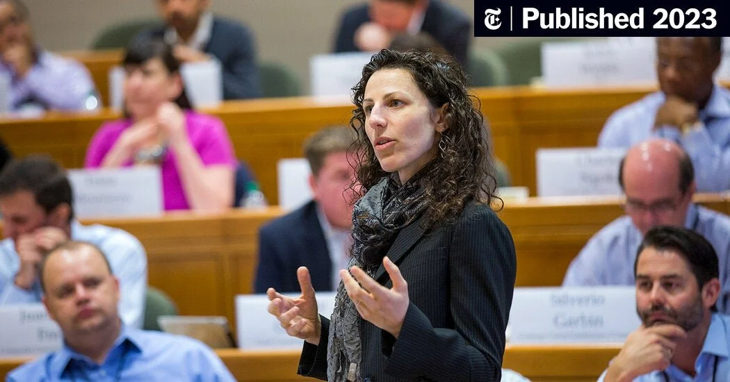 A woman with curly dark hair speaking at a conference with several seated attendees in the background.